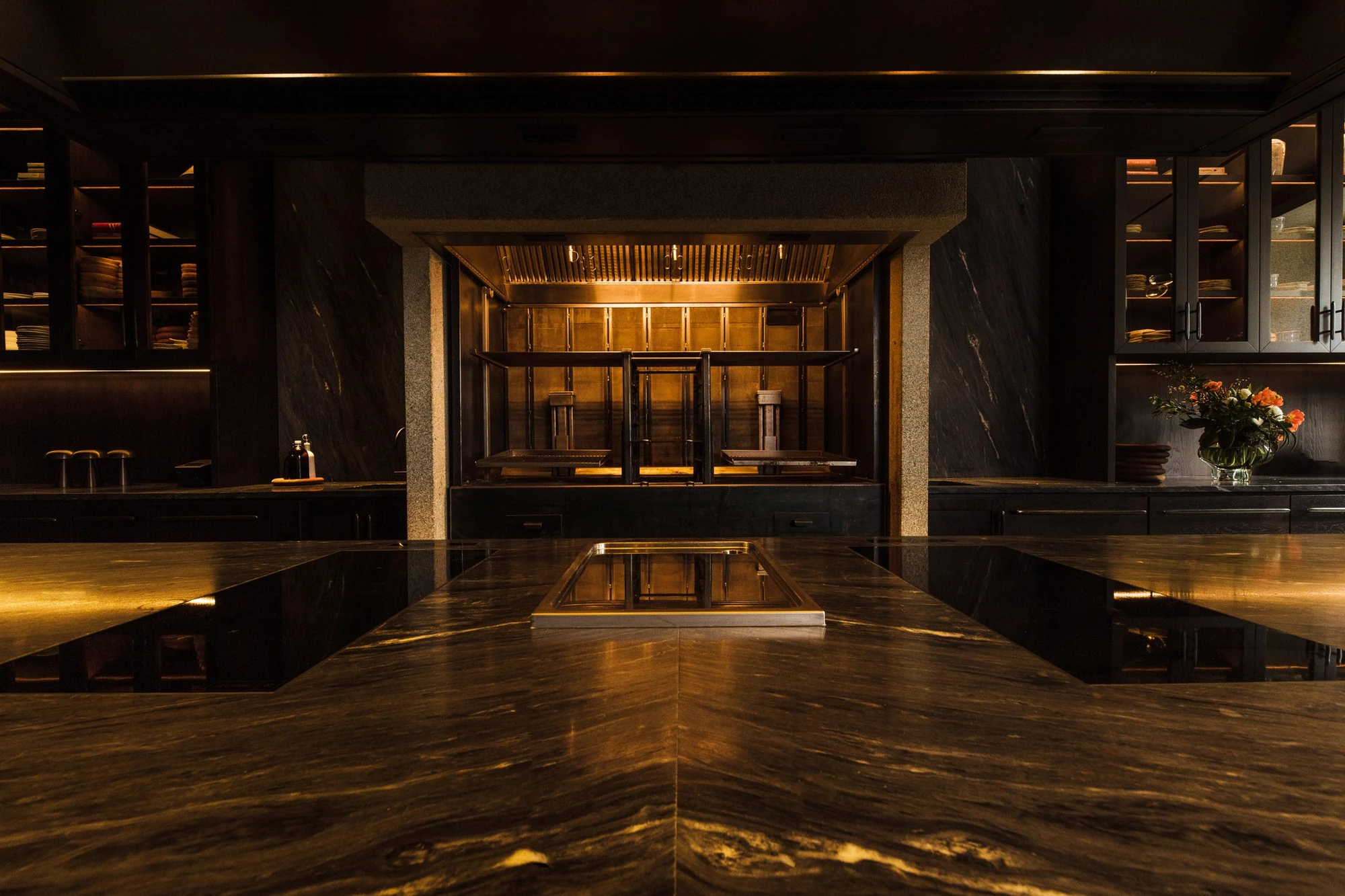Cozinha das Flores Mural by Álvaro Siza, photographed by Luís Moreira — The open kitchen at Cozinha das Flores seen across a veined stone pass, a brass-lined grill glowing between two granite piers.