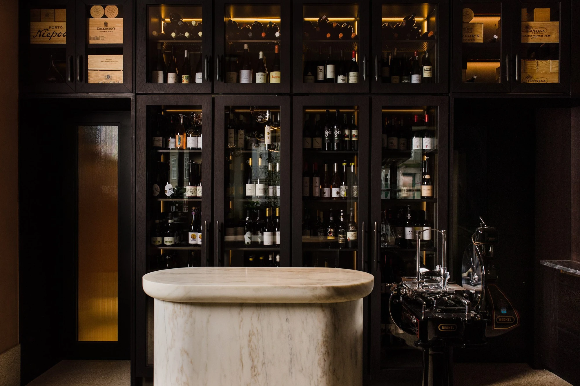 Cozinha das Flores Mural by Álvaro Siza, photographed by Luís Moreira — Oval white marble bar at Cozinha das Flores set against backlit cabinets of port and wine, with a chrome flywheel slicer at the right.