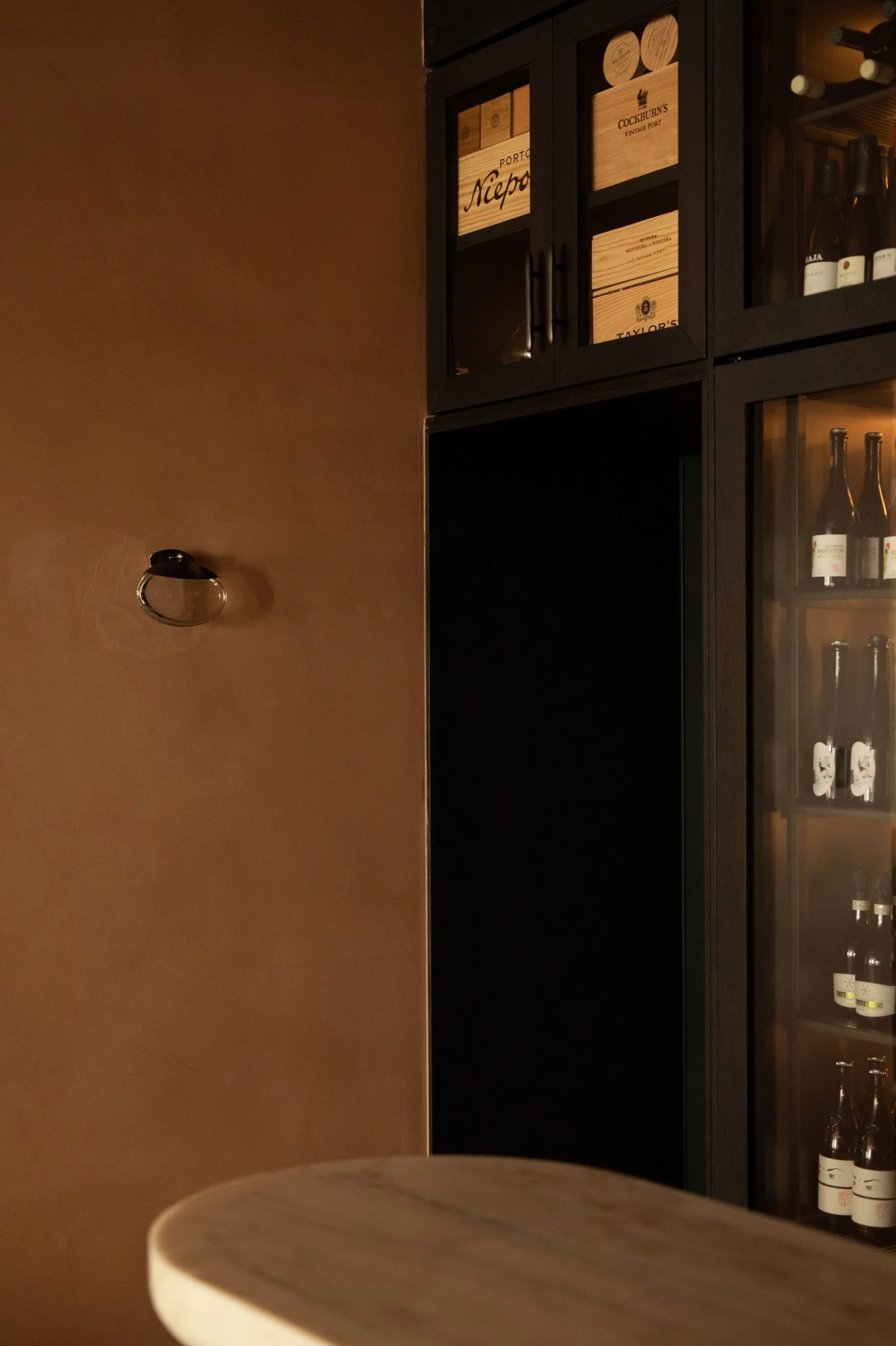 Cozinha das Flores Mural by Álvaro Siza, photographed by Luís Moreira — Coppery plastered wall and a brass door pull at Cozinha das Flores, beside dark cabinets stacked with wooden port cases.