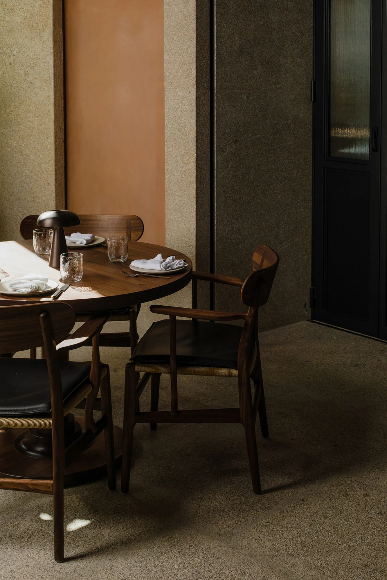 Cozinha das Flores Mural by Álvaro Siza, photographed by Luís Moreira — Round dark wood table and rush-seated chairs at Cozinha das Flores against a wall of coppery plaster and coarse granite.