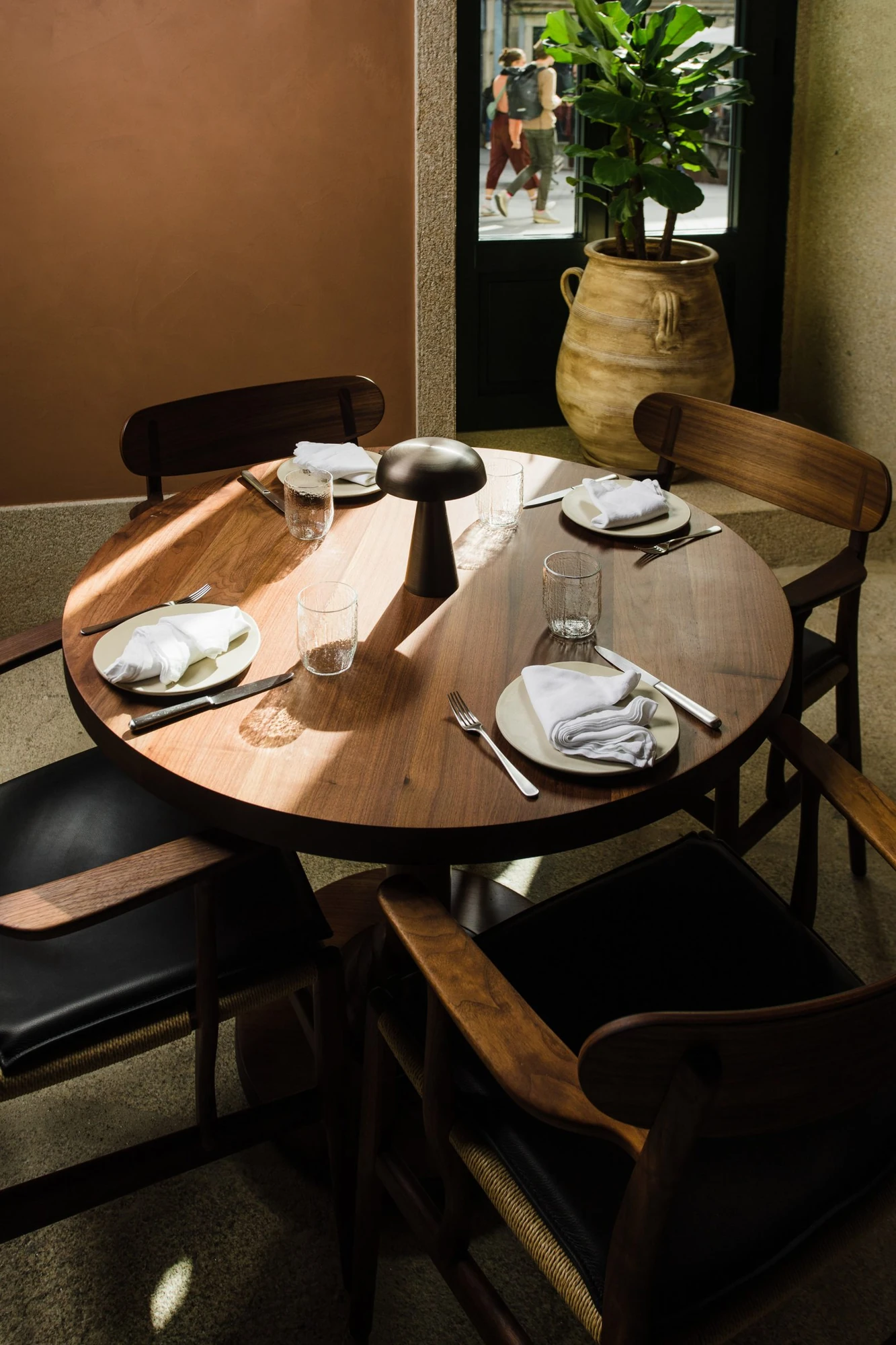 Cozinha das Flores Mural by Álvaro Siza, photographed by Luís Moreira — Corner table at Cozinha das Flores beside a green-framed door, with a terracotta urn, a fig-leafed plant and the street passing outside.