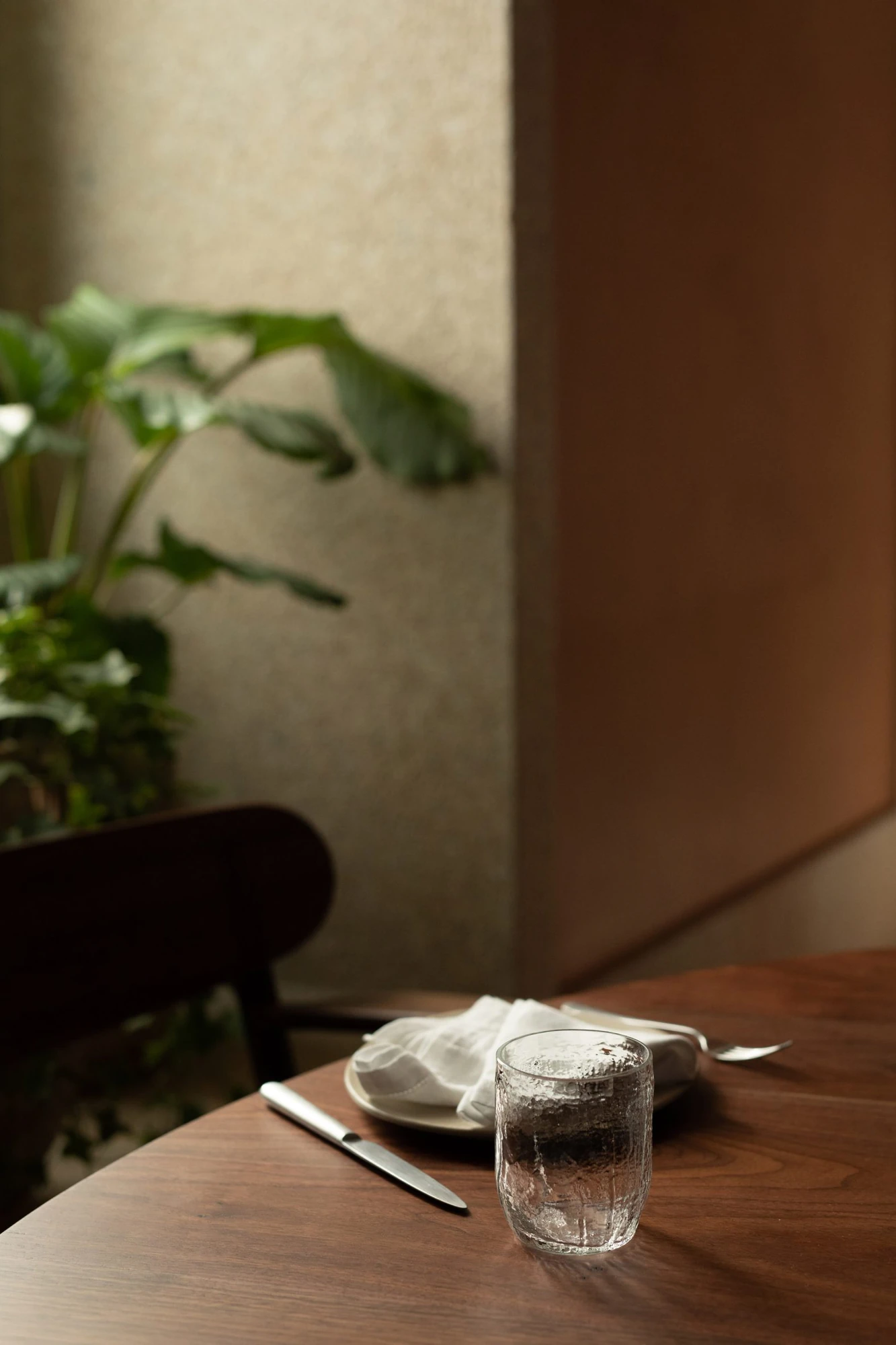 Cozinha das Flores Mural by Álvaro Siza, photographed by Luís Moreira — Hammered glass tumbler and folded napkin on a dark wood table at Cozinha das Flores, with foliage against plaster behind.
