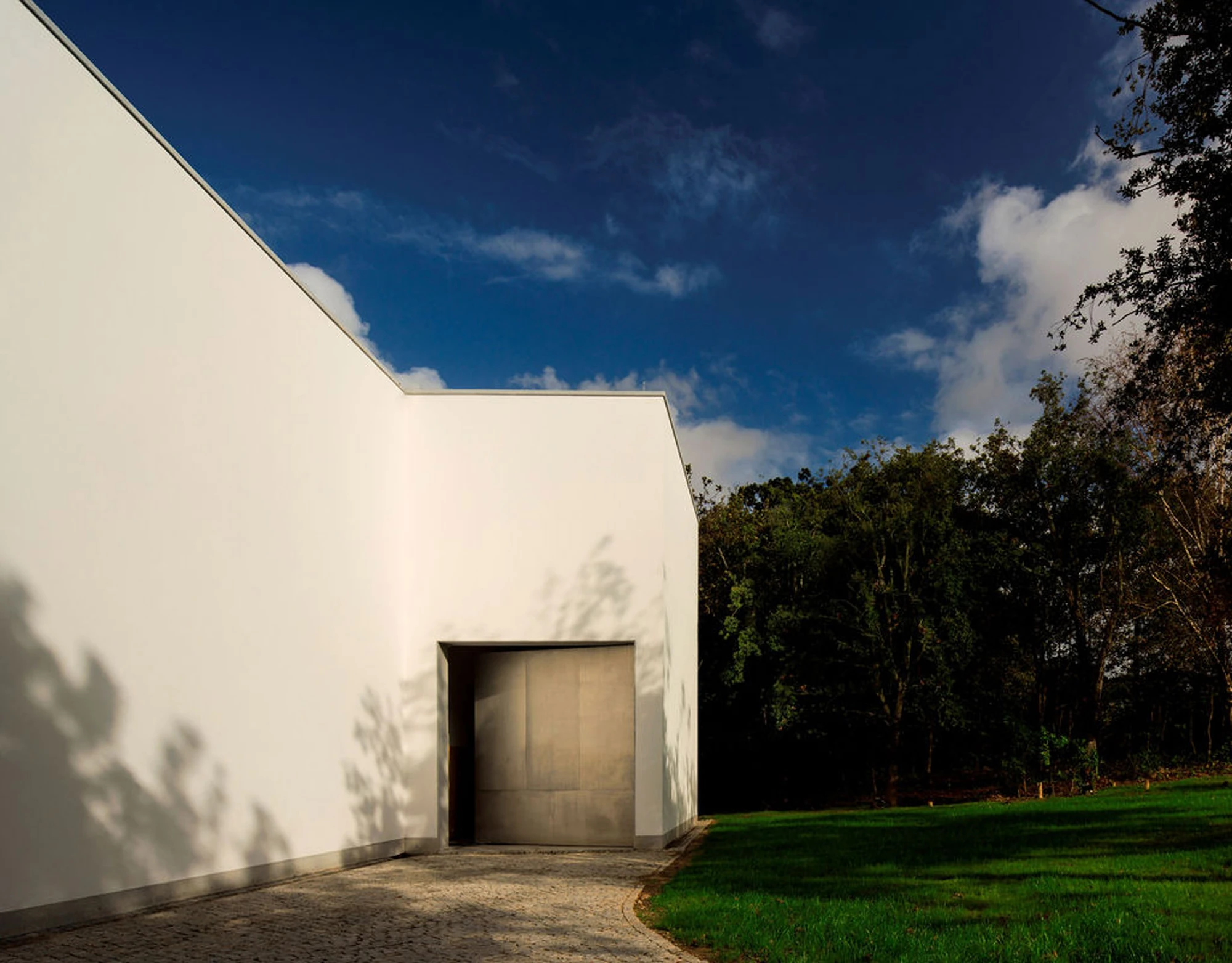 Serralves Museum New Wing by Álvaro Siza — White plastered wall of the Álvaro Siza Wing turning a corner to a recessed metal door, branch shadows falling across the plaster under a deep blue sky.