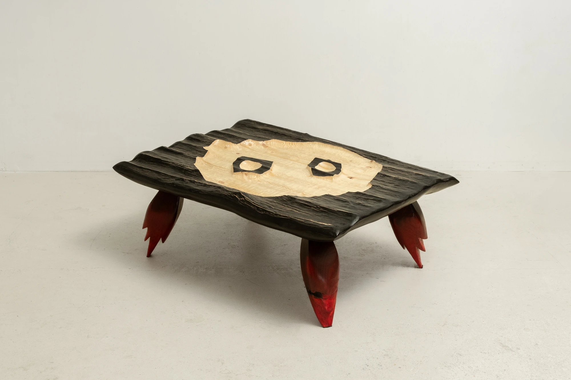 In Between Wood and Fire by Didi NG Wing Yin, photographed by Didi NG Wing Yin — The Rocket Side Coffee Table in full, its charred top cut back at the centre to a pale carved field holding two dark apertures, carried on four red legs that taper to points.