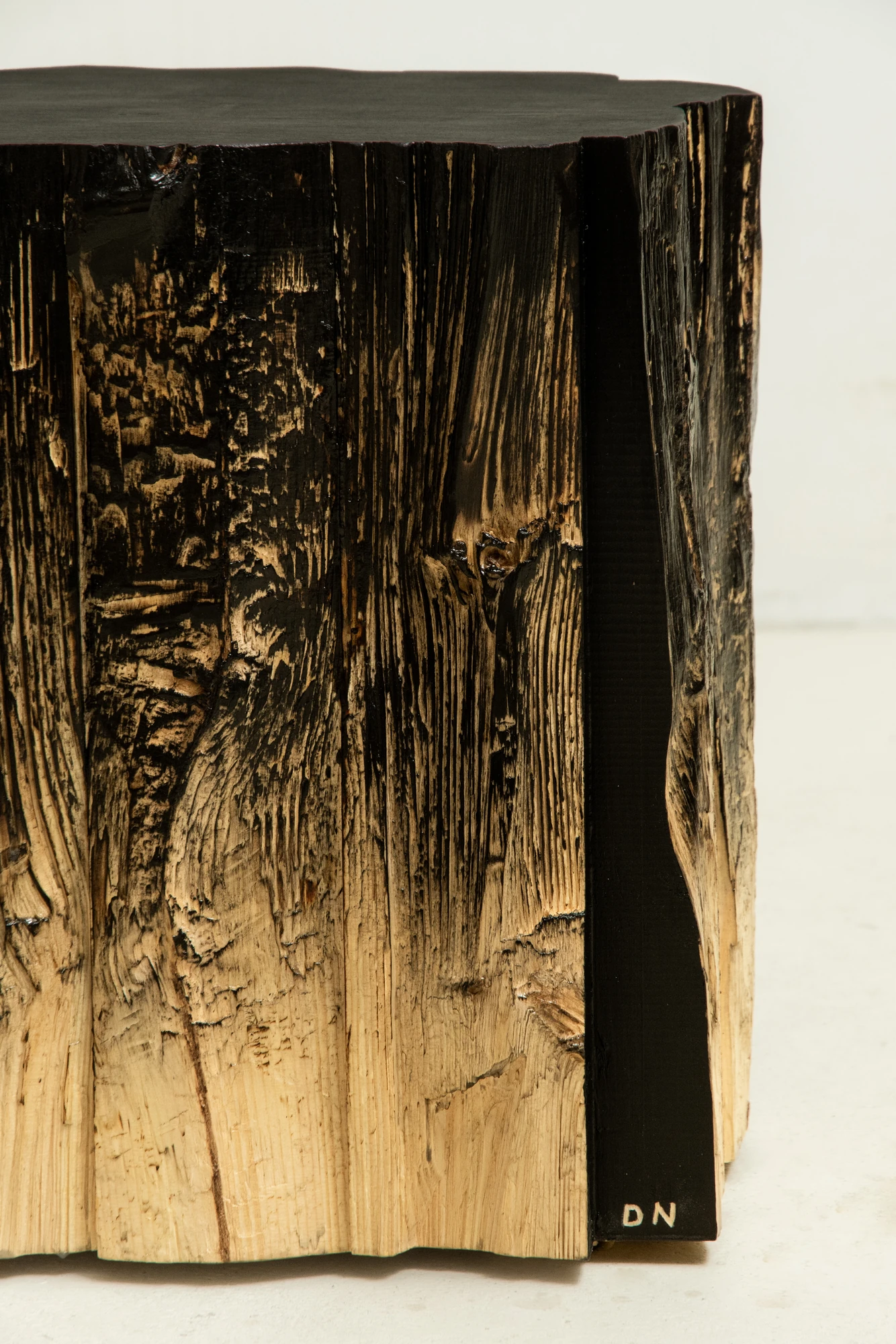 In Between Wood and Fire by Didi NG Wing Yin, photographed by Didi NG Wing Yin — Corner detail of a Pith stool where the char thins into bare fibre, with the initials DN lettered on the smooth black edge.