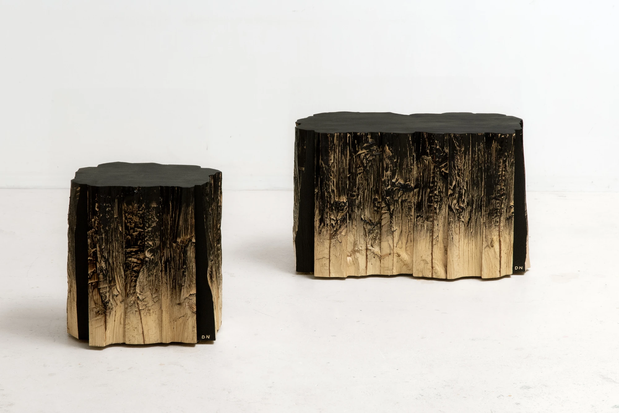 In Between Wood and Fire by Didi NG Wing Yin, photographed by Didi NG Wing Yin — Pith Stool A and Pith Stool B side by side, their split flanks running from near black at the seat down to raw pale timber at the foot.