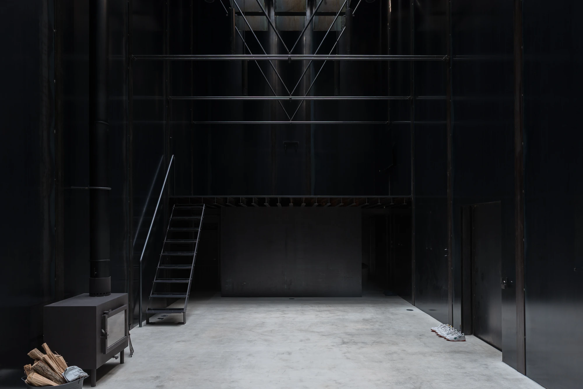 Shironishi Office by Tomoaki Uno Architects, photographed by Nathanael Bennett — The main room: black plate walls, a pale concrete floor, a wood stove at the left and a steel stair rising to the mezzanine.