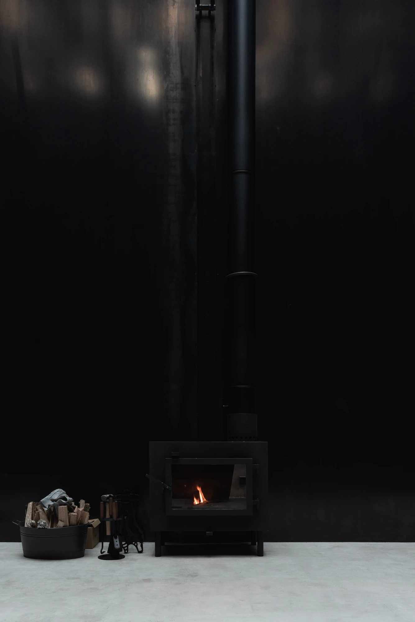Shironishi Office by Tomoaki Uno Architects, photographed by Nathanael Bennett — Fire lit in the stove, its flue running the full height of the black wall, logs and tools set on the concrete.
