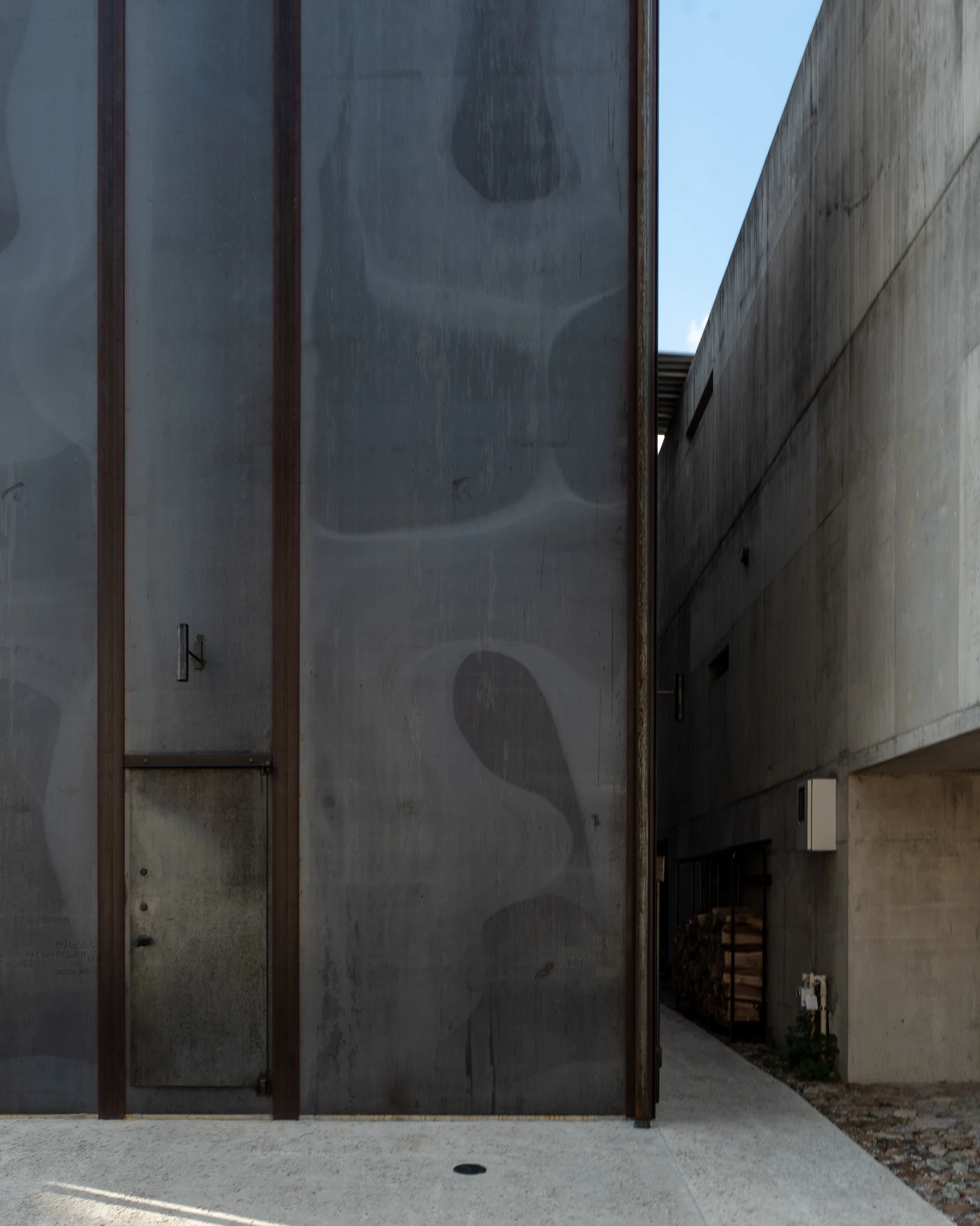 Shironishi Office by Tomoaki Uno Architects, photographed by Nathanael Bennett — The narrow gap between the steel wall and the concrete building next door, firewood stacked at the far end.