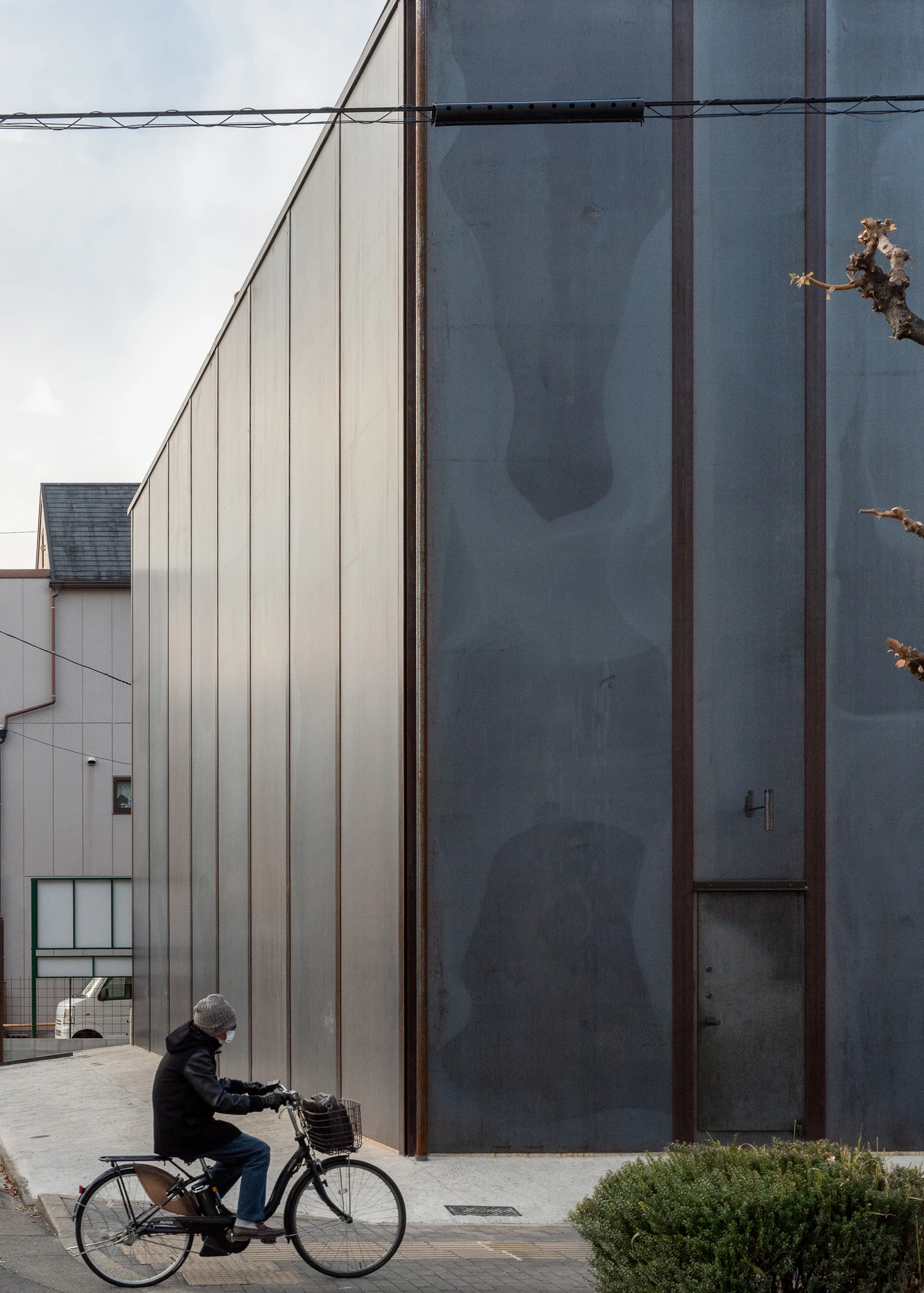 Shironishi Office by Tomoaki Uno Architects, photographed by Nathanael Bennett — Oblique view along the wall, one face catching low sun while a resident cycles past the flush service door.