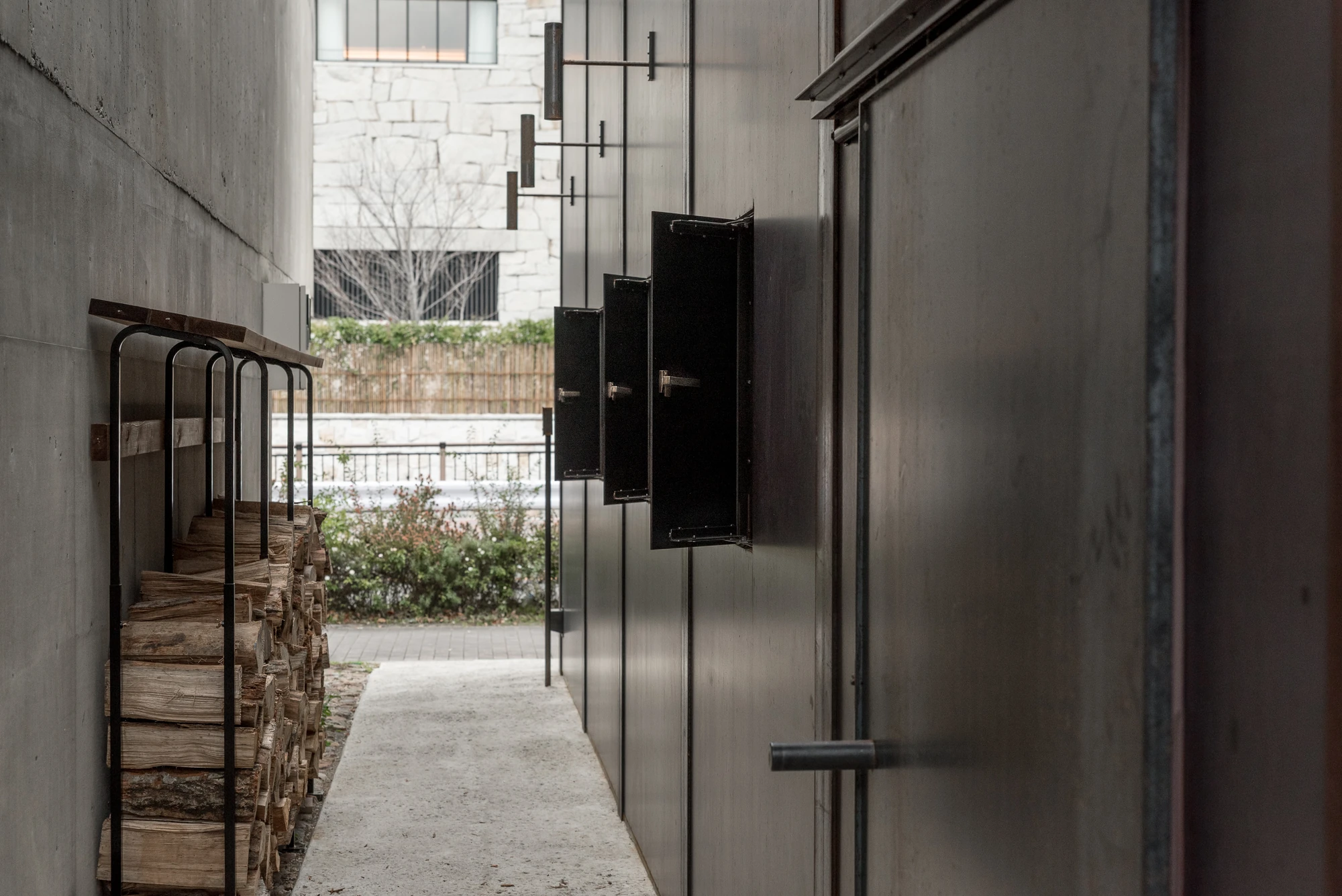 Shironishi Office by Tomoaki Uno Architects, photographed by Nathanael Bennett — Black steel hatches hinged open along the side wall above a rack of split logs, the alley opening onto a stone boundary wall.