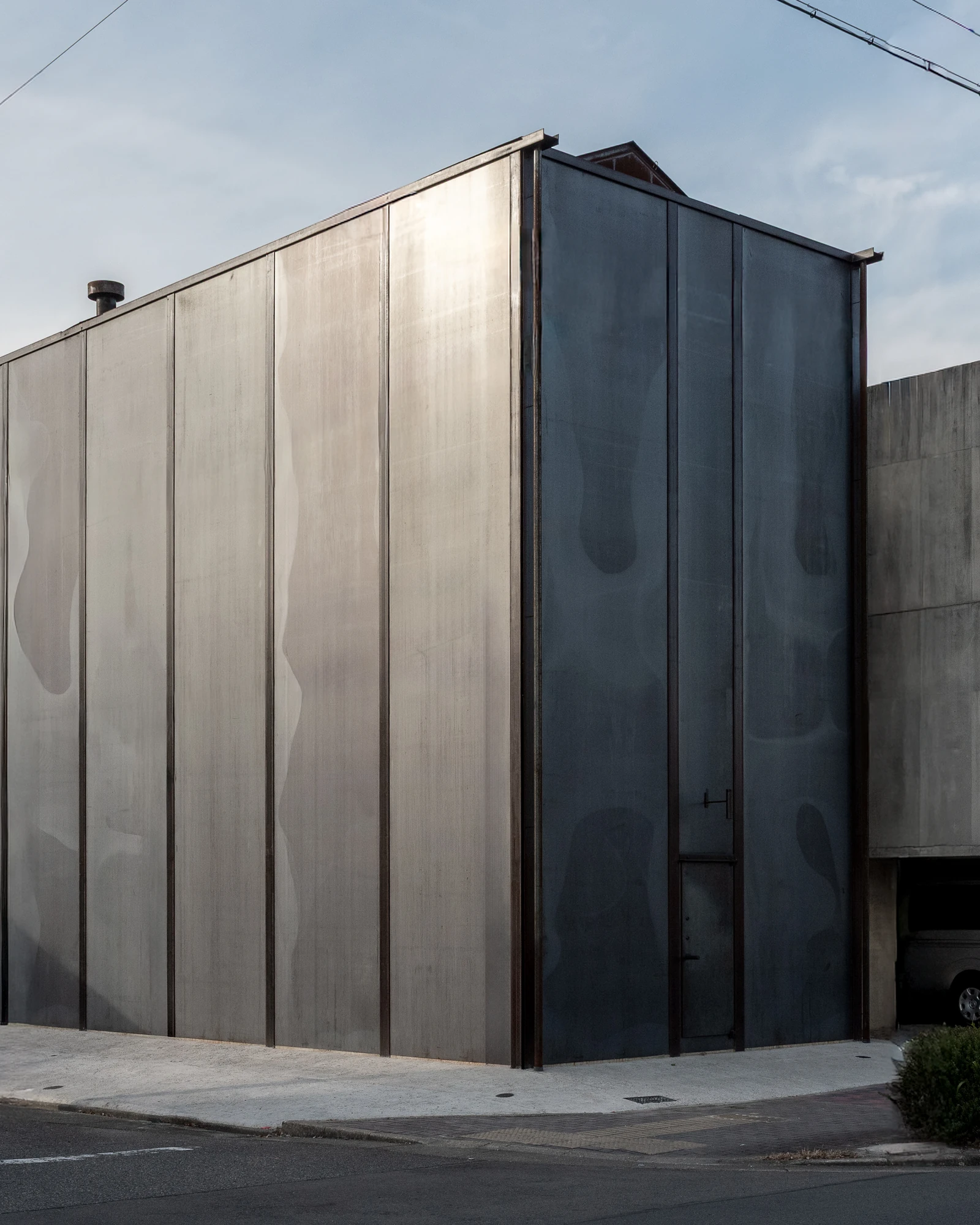 Shironishi Office by Tomoaki Uno Architects, photographed by Nathanael Bennett — The corner from the pavement, a silver-lit face turning into a slate-dark one, with a small plate door set into the base.