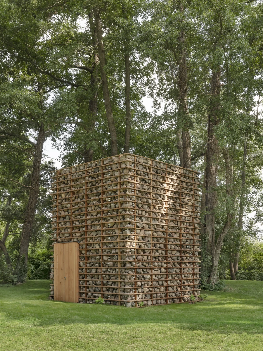 HAIN by undjurekbrüggen, photographed by Hannes Heitmüller — The fieldstone volume of HAIN standing on mown grass beneath tall poplars, horizontal larch battens holding loose grey stone across every face.