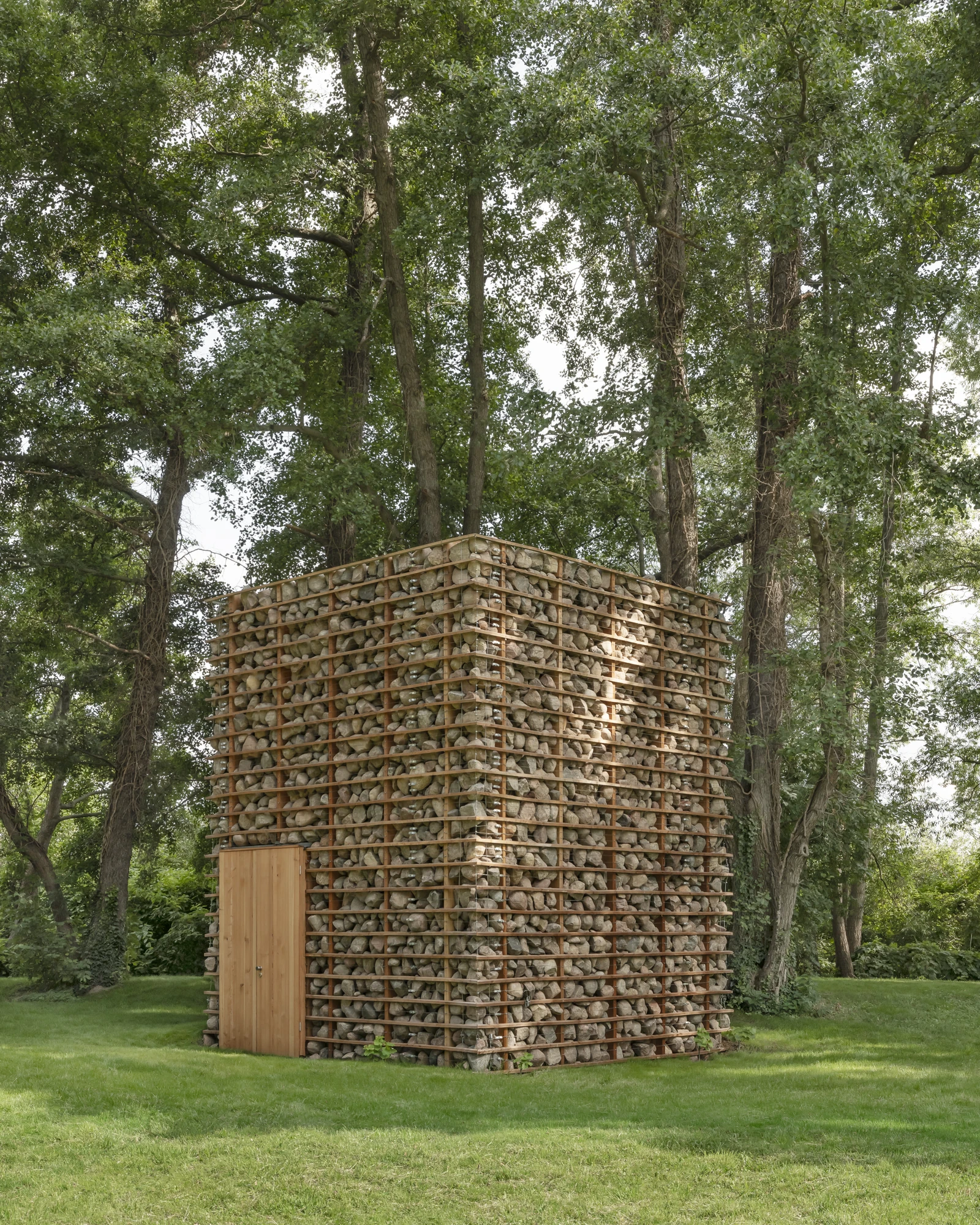 HAIN by undjurekbrüggen, photographed by Hannes Heitmüller — The fieldstone volume of HAIN standing on mown grass beneath tall poplars, horizontal larch battens holding loose grey stone across every face.