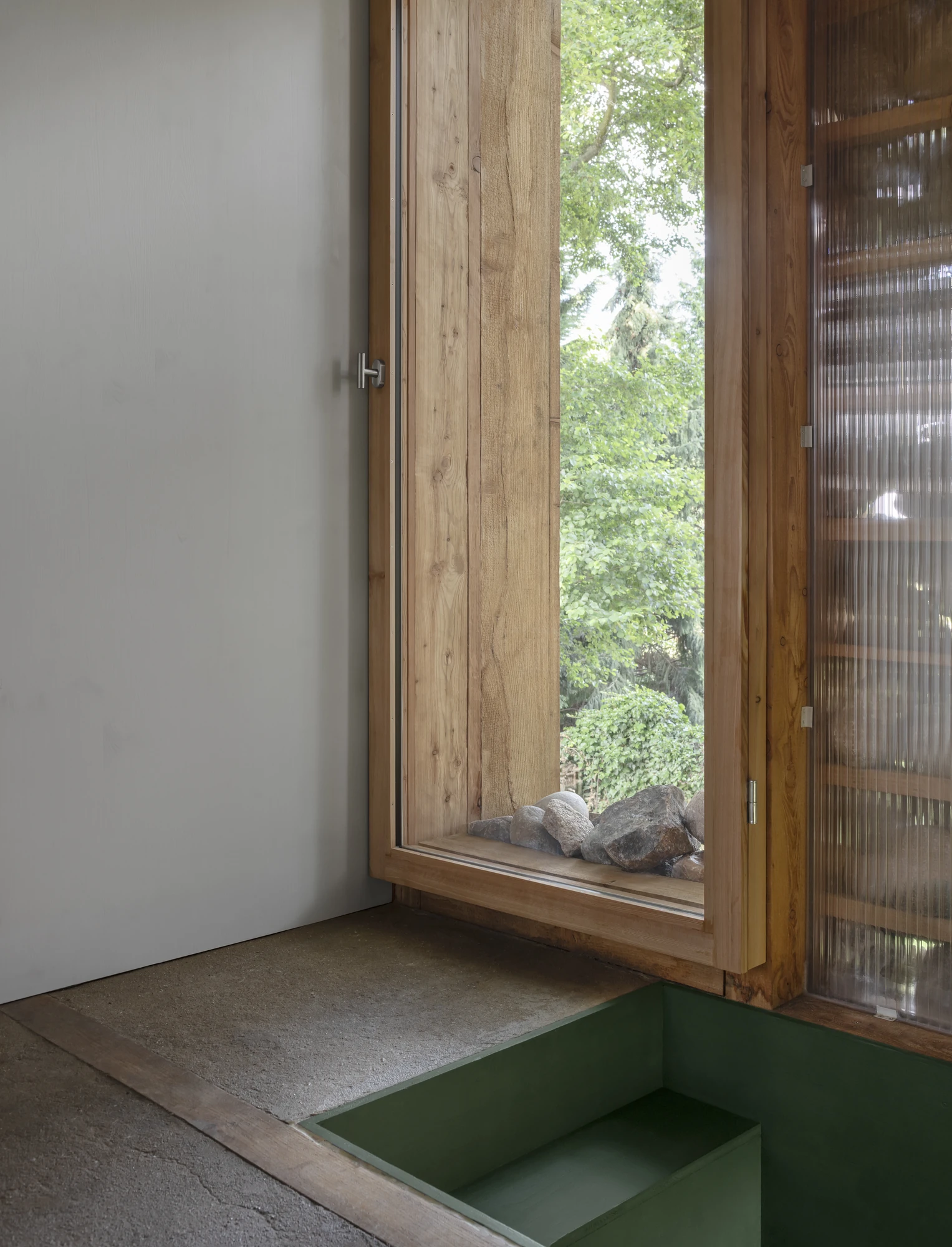 HAIN by undjurekbrüggen, photographed by Hannes Heitmüller — An interior corner where a timber window opens onto trees, stones resting on the sill and a green-lined recess cut into the floor.
