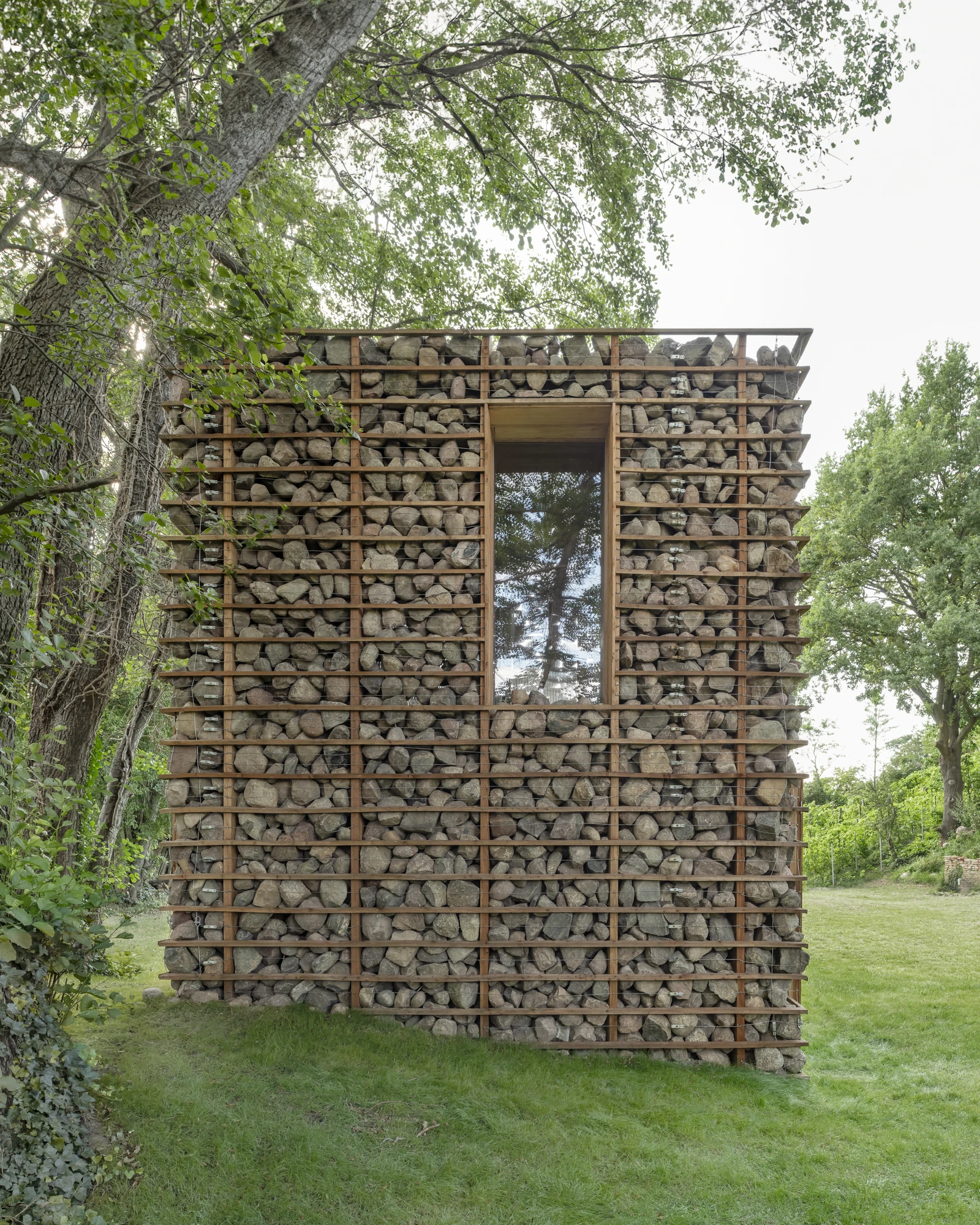 HAIN by undjurekbrüggen, photographed by Hannes Heitmüller — The stone facade seen straight on, a tall narrow window slot cut through the layered fieldstone and lined in pale timber.