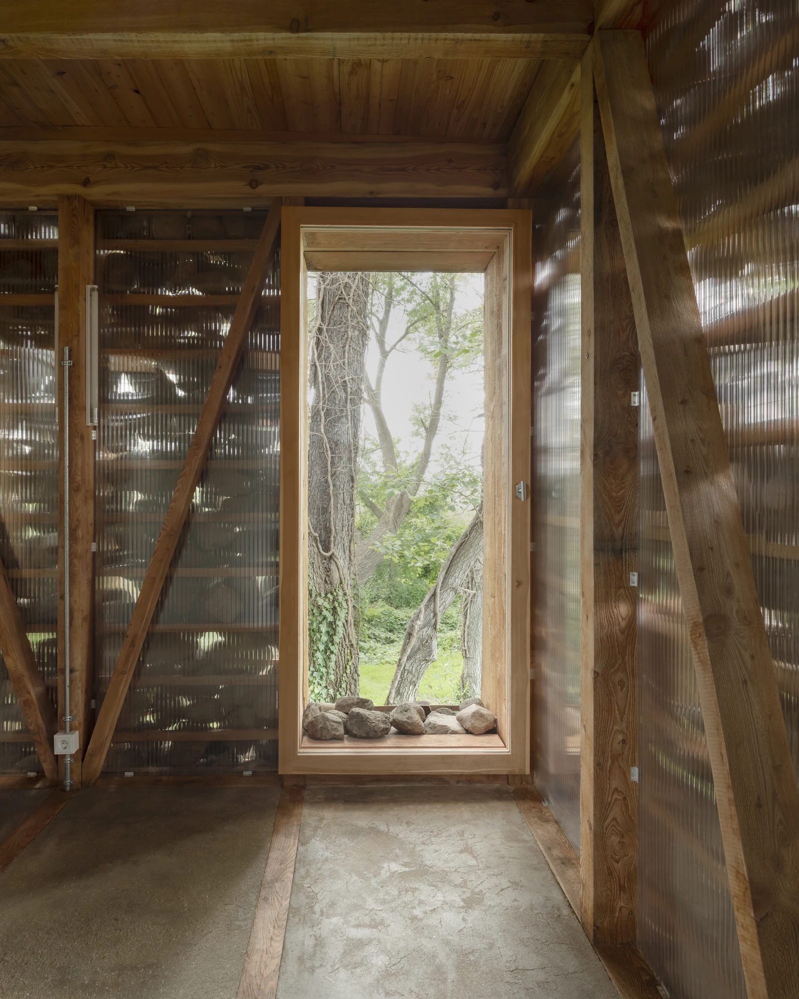 HAIN by undjurekbrüggen, photographed by Hannes Heitmüller — A larch-framed window set into a wall of translucent corrugated panels, fieldstones laid along the sill and a screed floor underfoot.
