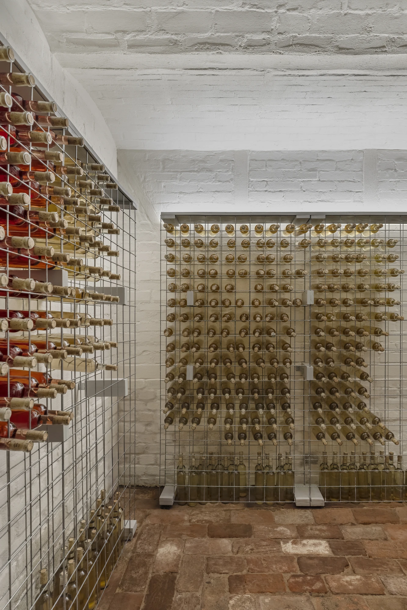 HAIN by undjurekbrüggen, photographed by Hannes Heitmüller — The cellar below, wine bottles racked in steel mesh against whitewashed brick, with a shallow cap vault overhead and worn brick pavers underfoot.