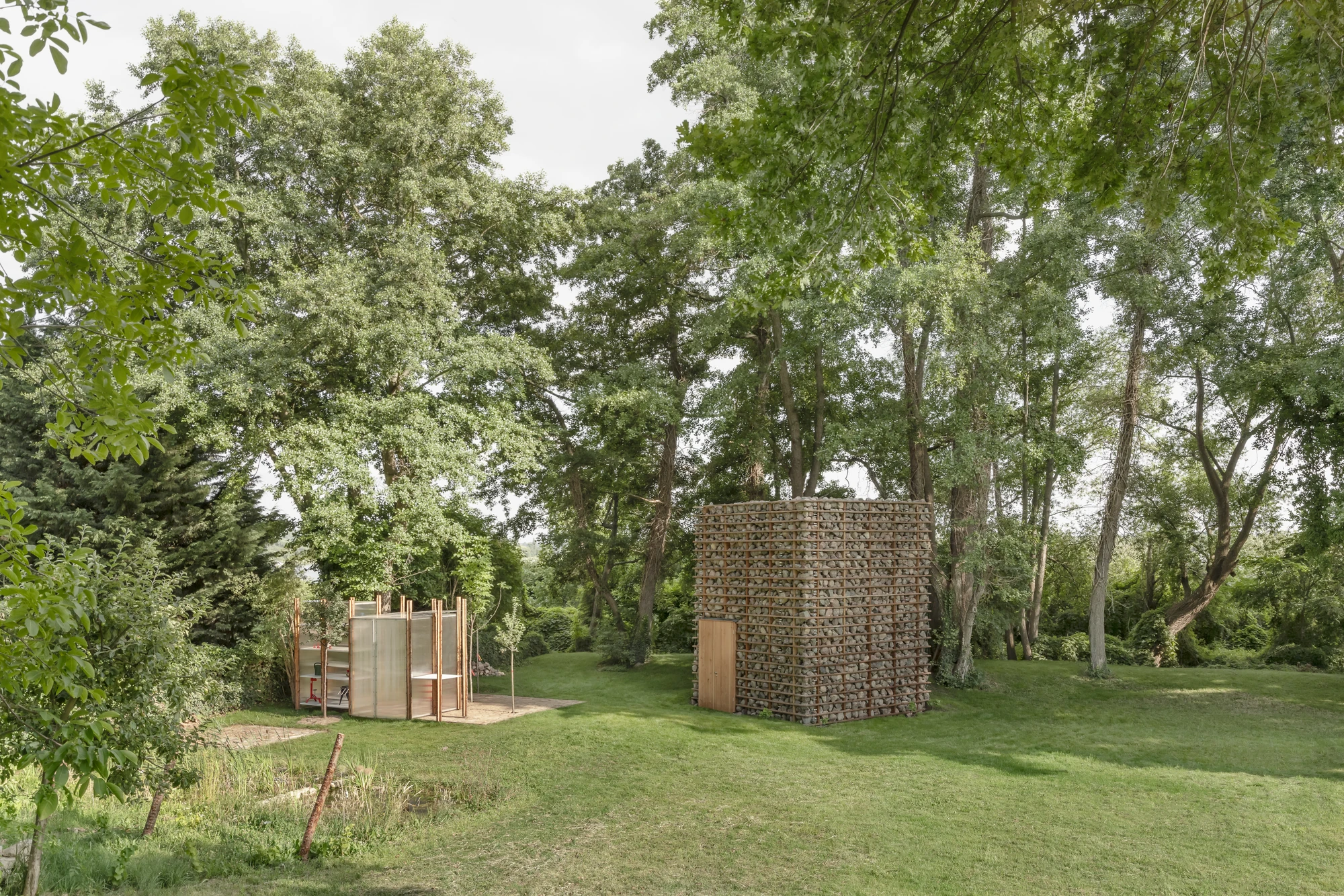 HAIN by undjurekbrüggen, photographed by Hannes Heitmüller — Both structures across open lawn: the stone volume among mature trees and the translucent pavilion set on a brick paved terrace.