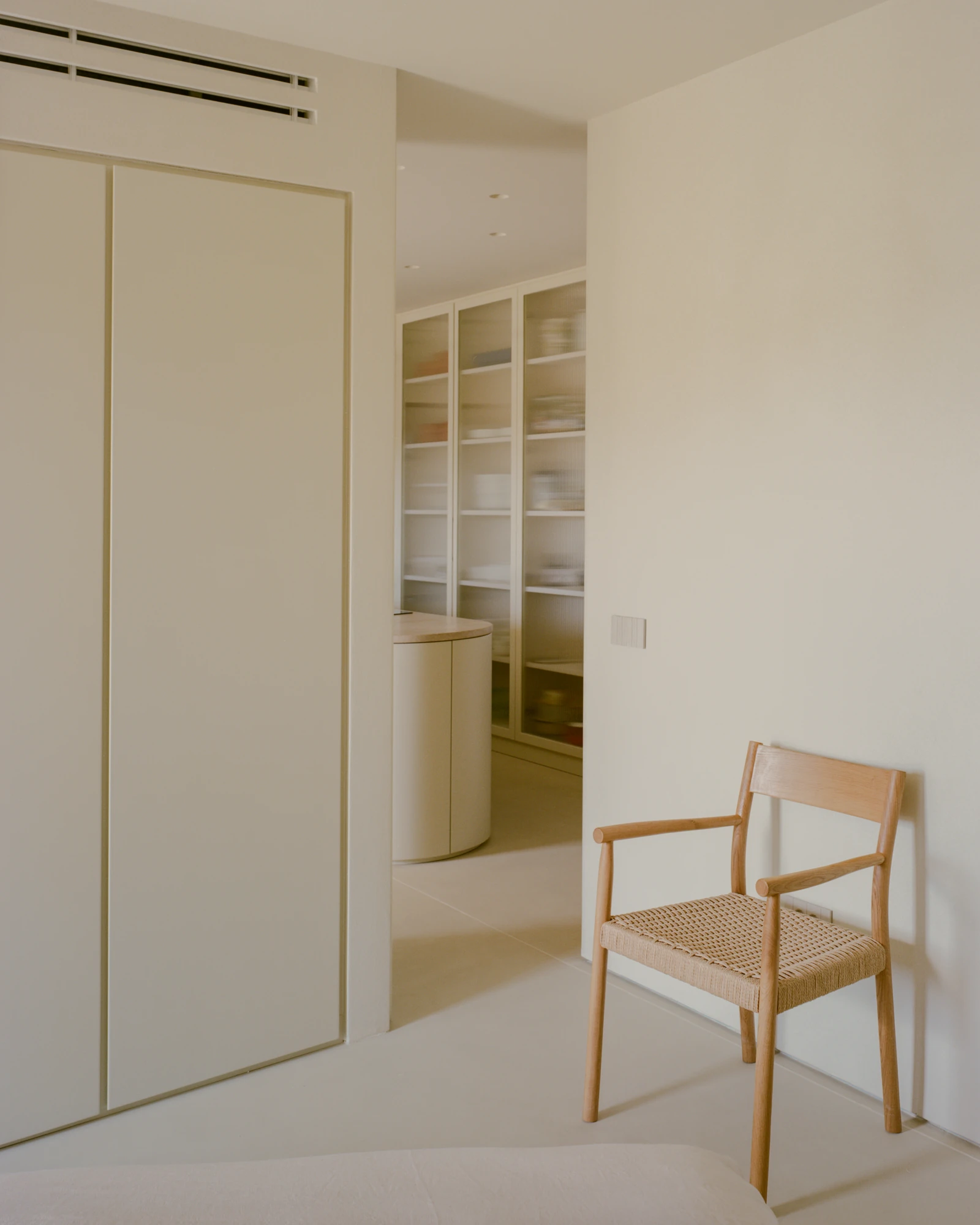 Kifisia Apartment by Georgios Apostolopoulos Architects, photographed by Lorenzo Zandri — A flush wardrobe front beside an opening back toward the kitchen island, with a caned oak armchair against the wall.