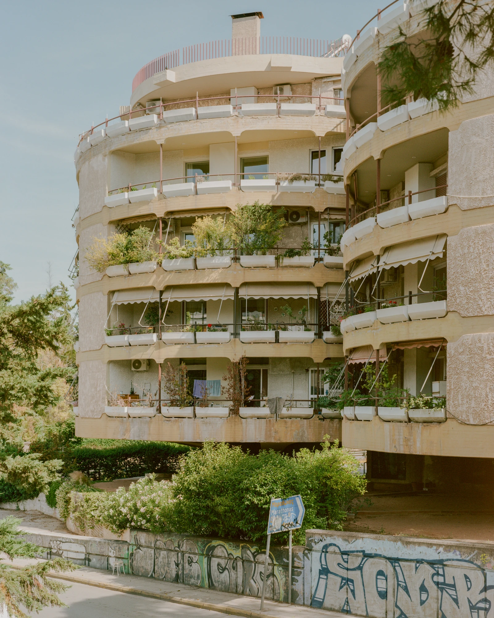 Kifisia Apartment by Georgios Apostolopoulos Architects, photographed by Lorenzo Zandri — The curved 1970s block from the street, its stacked concrete balcony trays overgrown with decades of resident planting.