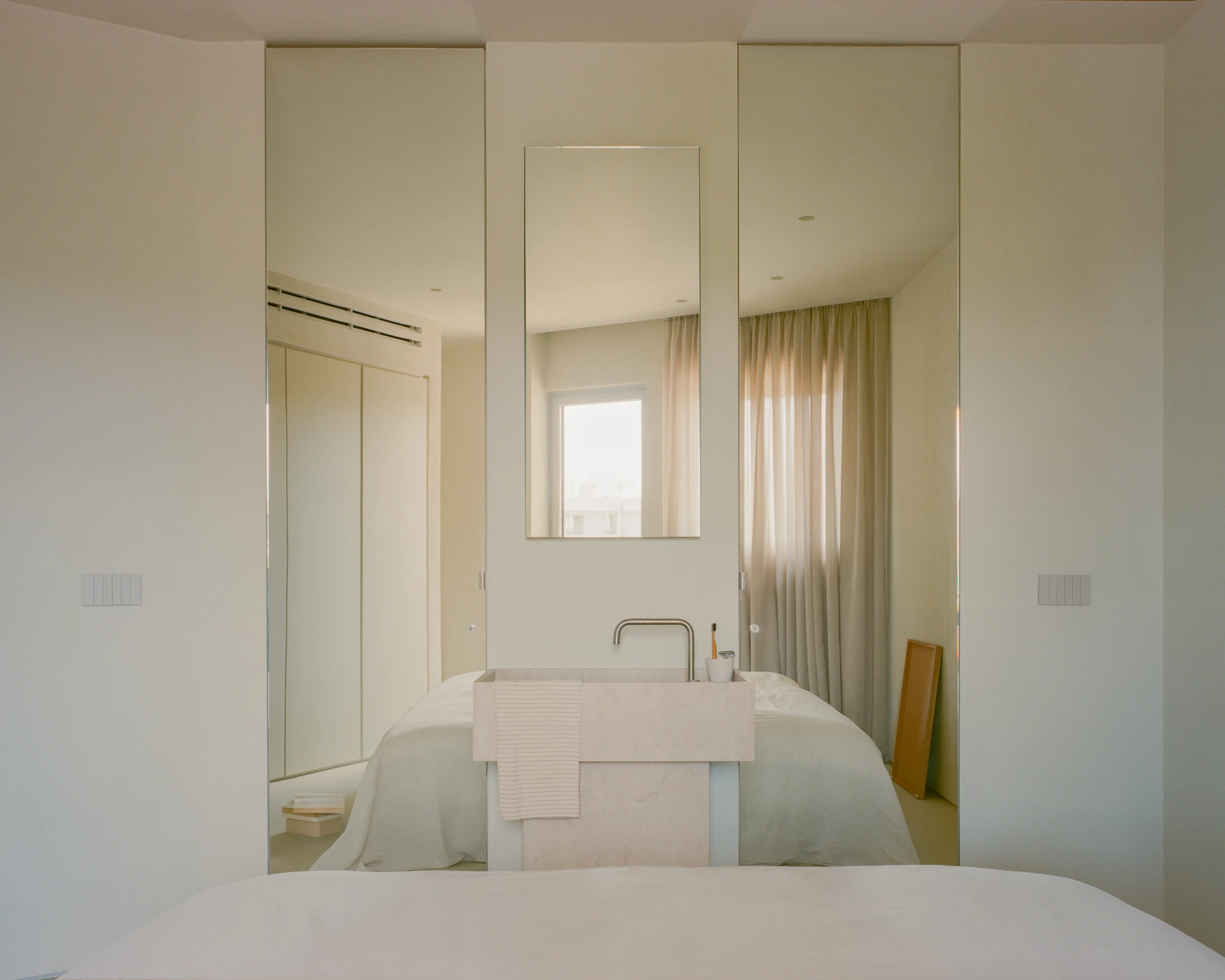 Kifisia Apartment by Georgios Apostolopoulos Architects, photographed by Lorenzo Zandri — The pale stone vanity standing clear of the bathroom enclosure, mirrored panels doubling the bed and the plant beside it.