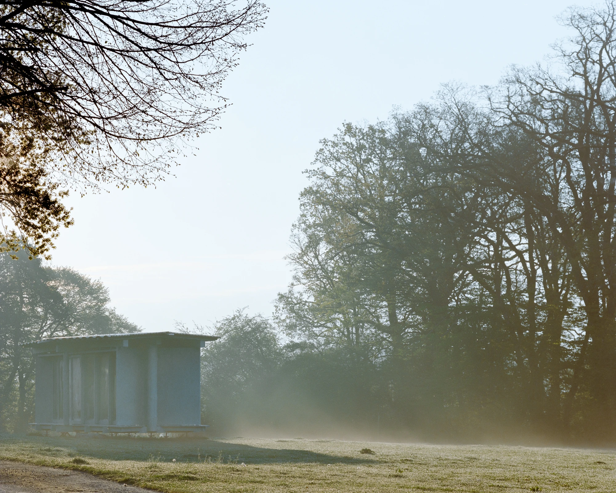 Pavilion of the Sliding Doors by Piertzovanis Toews, photographed by Federico Farinatti — Ground mist crosses the meadow at dawn and the pavilion sits small and blue grey at the left edge of the frame.