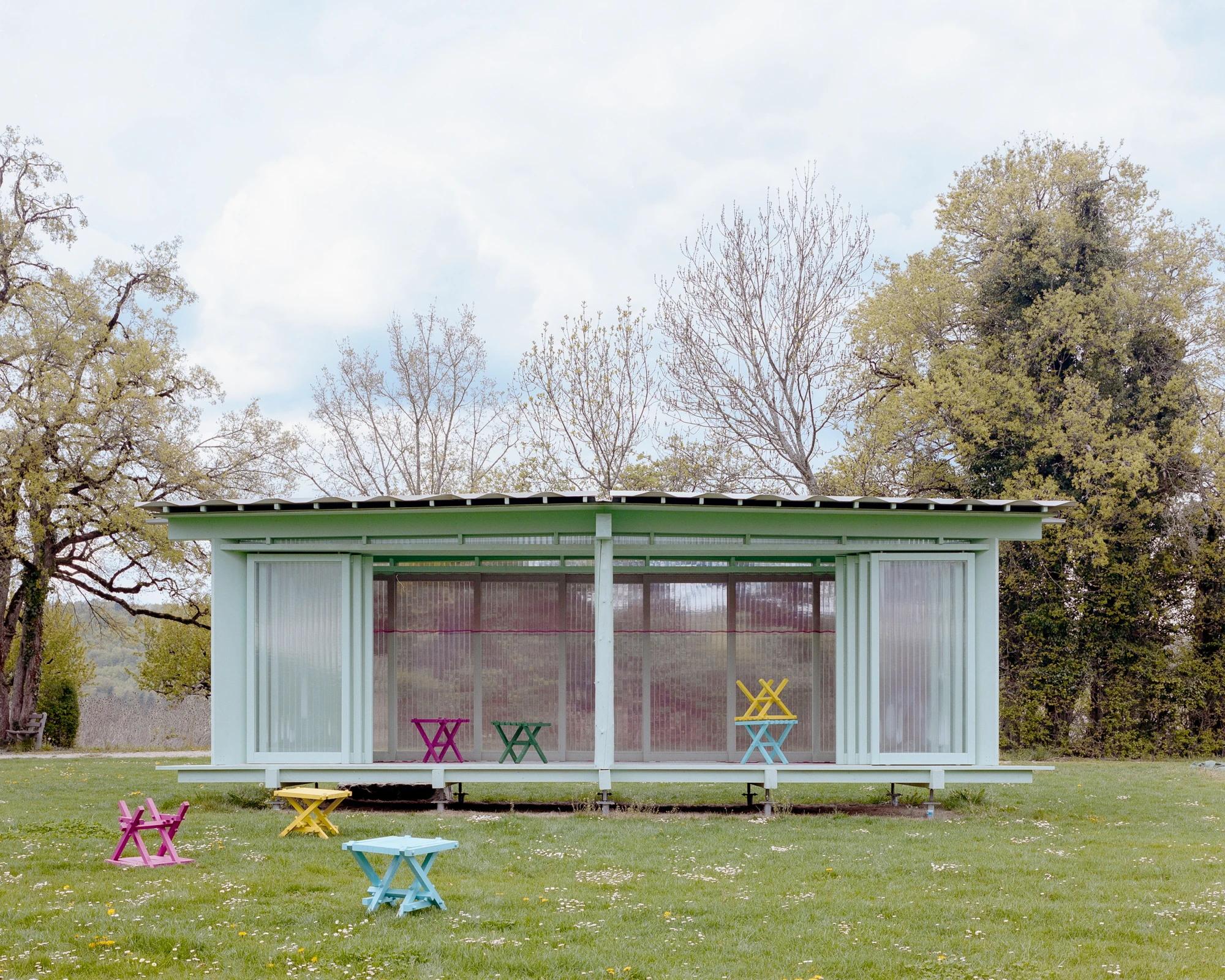 Pavilion of the Sliding Doors by Piertzovanis Toews, photographed by Federico Farinatti — The pavilion seen square on under a cloudy sky, with pink and yellow and blue folding stools scattered inside and out on the meadow.