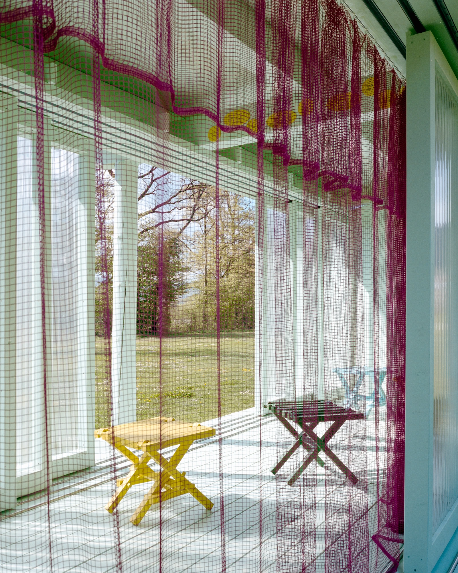 Pavilion of the Sliding Doors by Piertzovanis Toews, photographed by Federico Farinatti — Looking along the inside of the pavilion through a magenta mesh curtain, with yellow and dark red folding stools on the white floor.