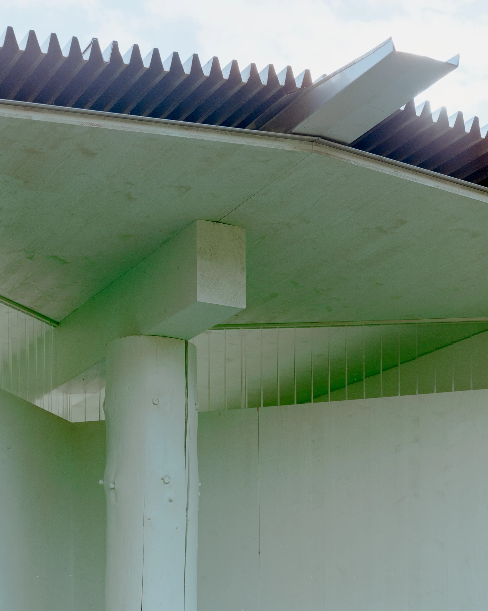 Pavilion of the Sliding Doors by Piertzovanis Toews, photographed by Federico Farinatti — Looking up at the corner of the roof, where dark corrugated metal sheets project past the pale green eaves.