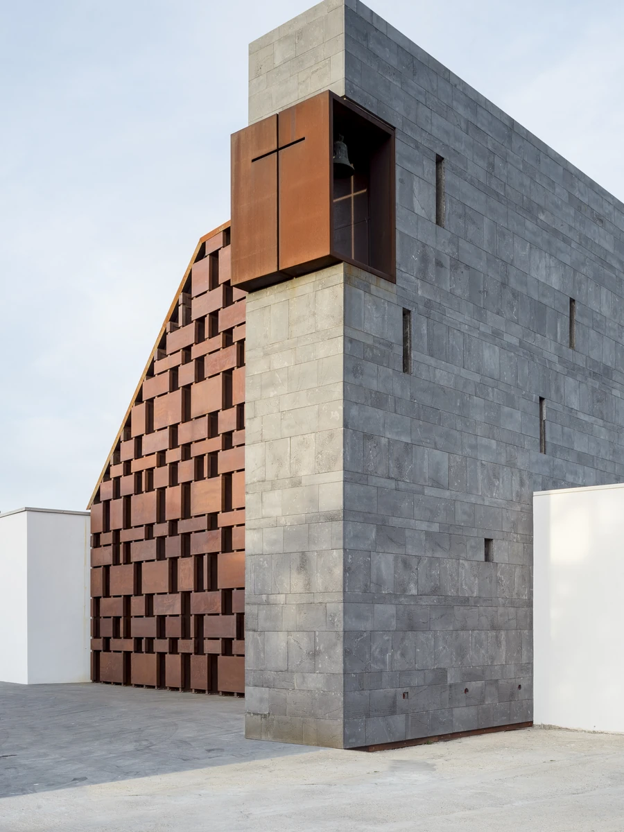 Complesso parrocchiale e Chiesa della Beata Vergine Assunta by Carlo Atzeni, Silvia Mocci, Maurizio Manias, Franceschino Serra, photographed by Studio Vetroblu — The corner of the dark basalt volume, with a bronze bell hung inside a corten steel opening…