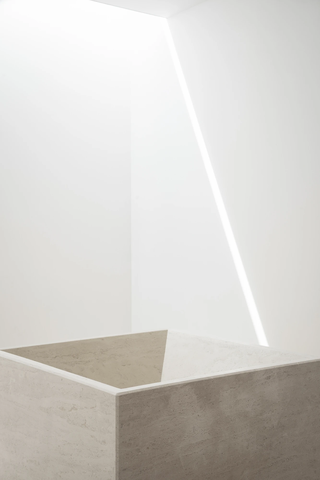 Complesso parrocchiale e Chiesa della Beata Vergine Assunta by Carlo Atzeni, Silvia Mocci, Maurizio Manias, Franceschino Serra, photographed by Studio Vetroblu — An angled slot of skylight draws a line of light down the white wall above a pale marble…