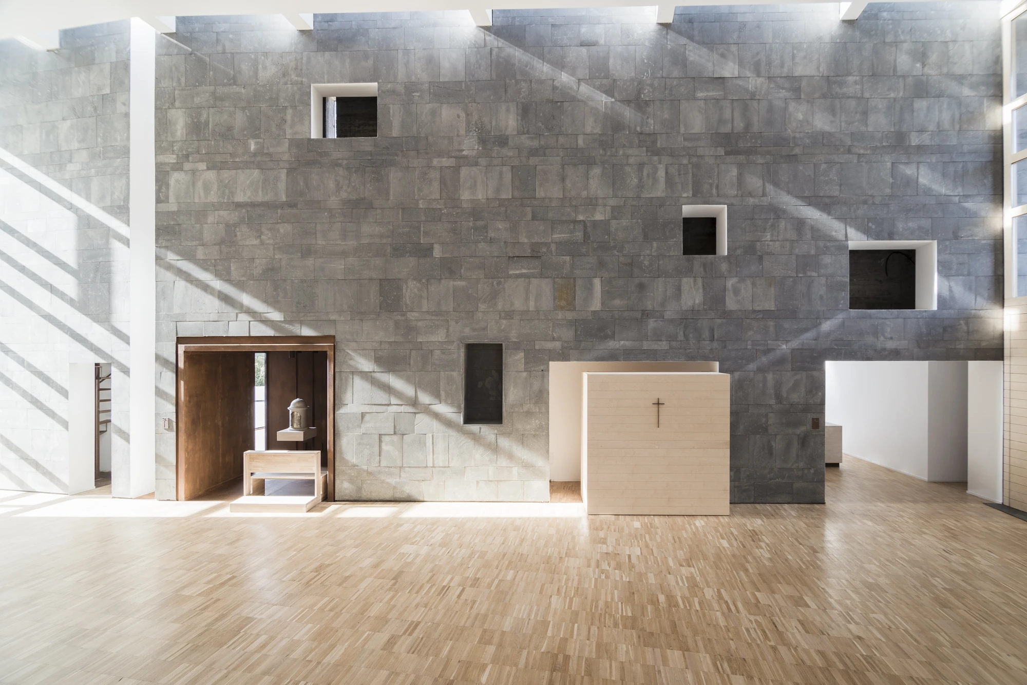 Complesso parrocchiale e Chiesa della Beata Vergine Assunta by Carlo Atzeni, Silvia Mocci, Maurizio Manias, Franceschino Serra, photographed by Studio Vetroblu — The full width of the basalt wall inside the hall, its rectangular openings scattered…
