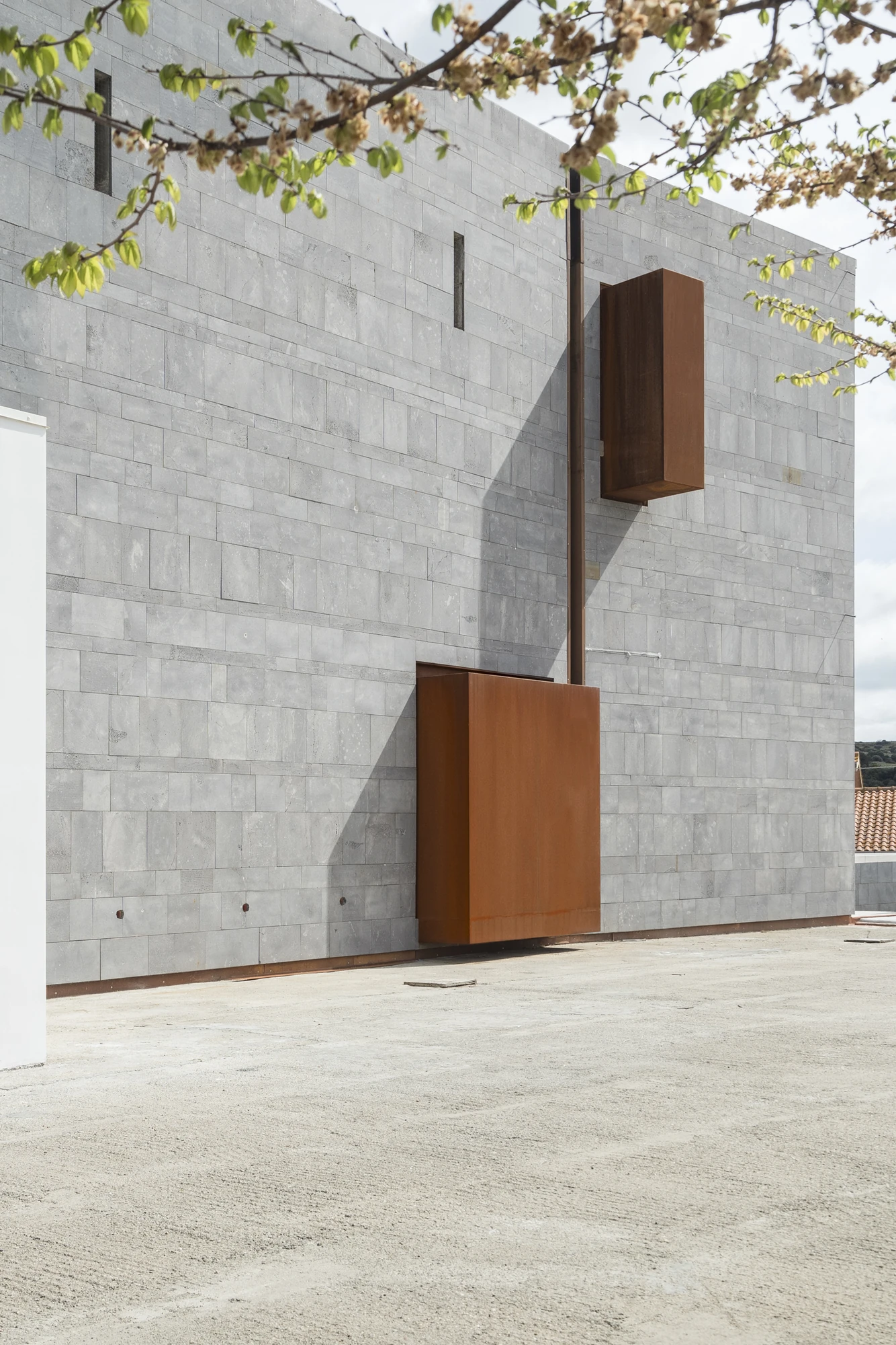 Complesso parrocchiale e Chiesa della Beata Vergine Assunta by Carlo Atzeni, Silvia Mocci, Maurizio Manias, Franceschino Serra, photographed by Studio Vetroblu — Late sun rakes the coursed basalt of the wall, seen past a flowering branch.