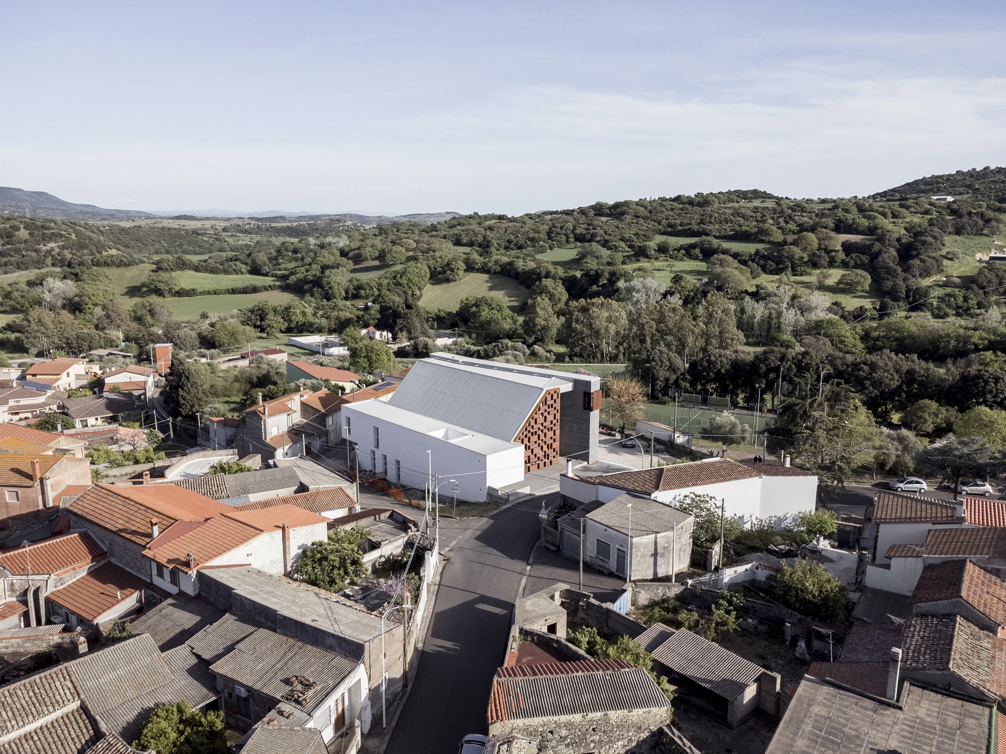 Complesso parrocchiale e Chiesa della Beata Vergine Assunta by Carlo Atzeni, Silvia Mocci, Maurizio Manias, Franceschino Serra, photographed by Studio Vetroblu — Seen from the air, the white church volume and its seamed metal roof sit among the red…