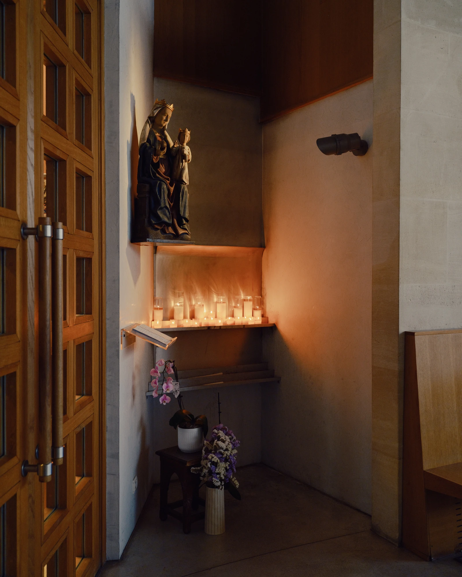 Église Saint-François de Molitor by Corinne Callies and Jean-Marie Duthilleul — A corner chapel lit by a single lamp, with a polychrome carved Virgin and Child on a stone bracket above a shelf of votive candles.