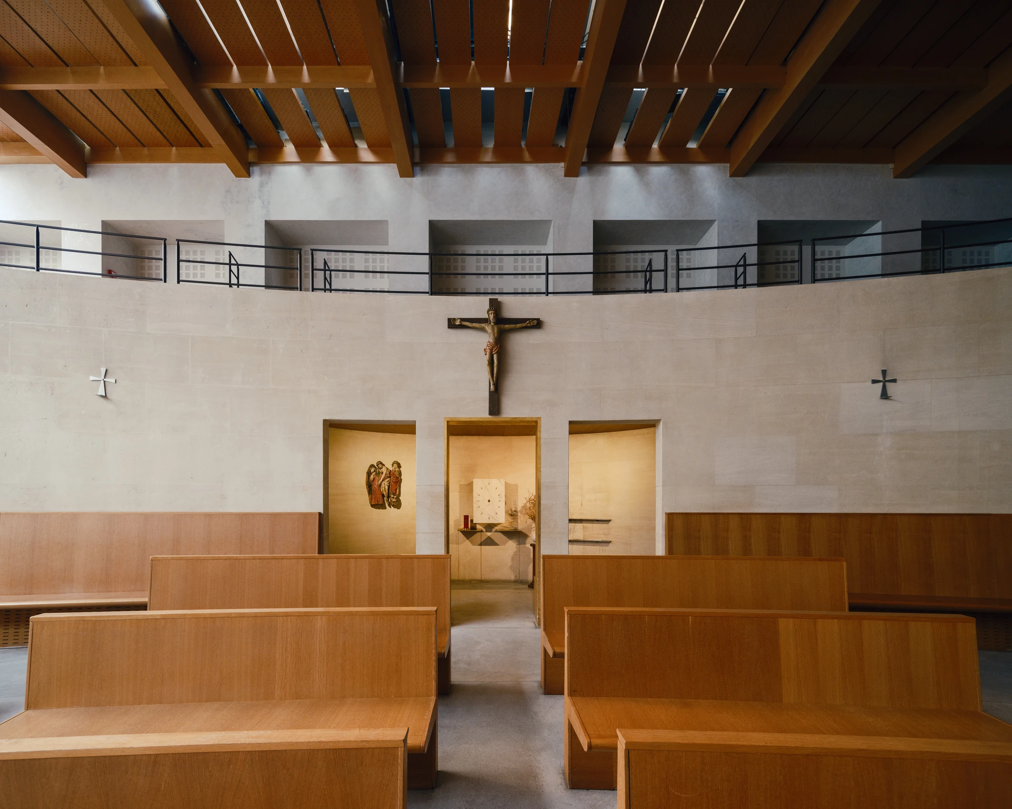 Église Saint-François de Molitor by Corinne Callies and Jean-Marie Duthilleul — A symmetrical view across empty timber benches towards a pale stone wall carrying a carved crucifix, flanked by two small metal crosses.