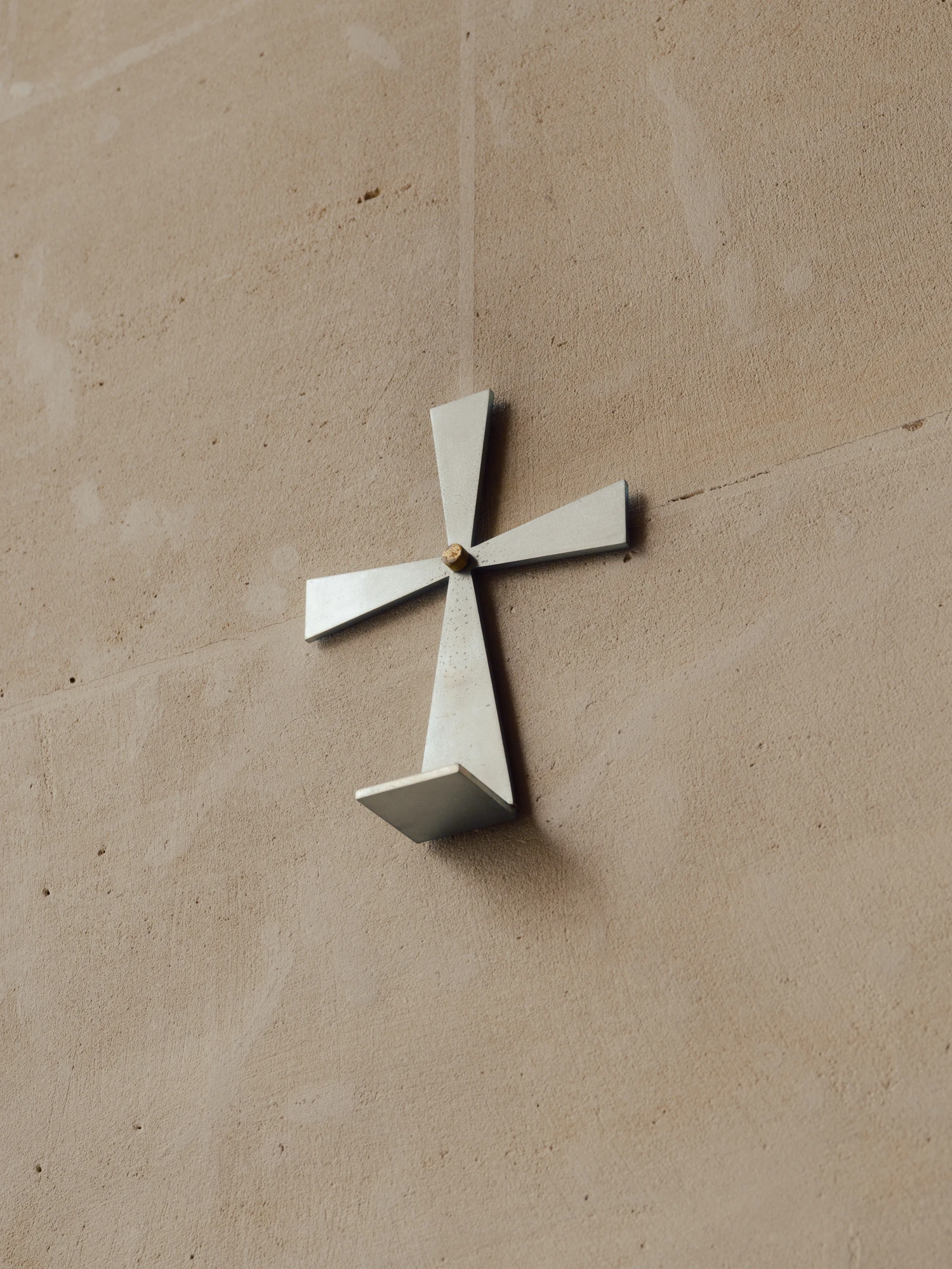 Église Saint-François de Molitor by Corinne Callies and Jean-Marie Duthilleul — A folded metal cross fixed to a pale plaster wall, its four arms angled so that each face catches the light differently.
