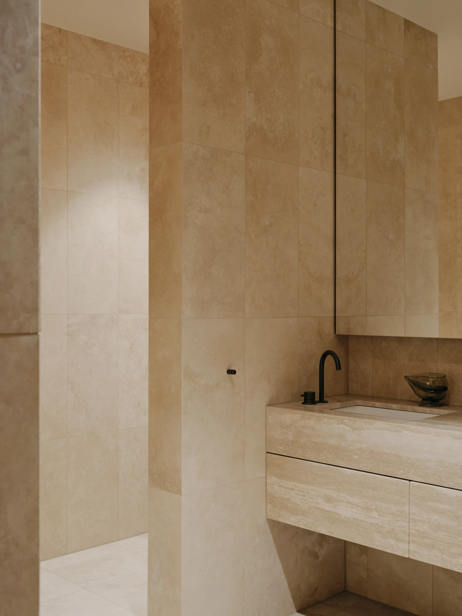 49 Walsh Street by MORQ, photographed by Tom Ross — A travertine bathroom seen past a partition wall, with a wall-hung stone vanity, black tapware, a dark glass bowl and a full-height mirrored cabinet above.