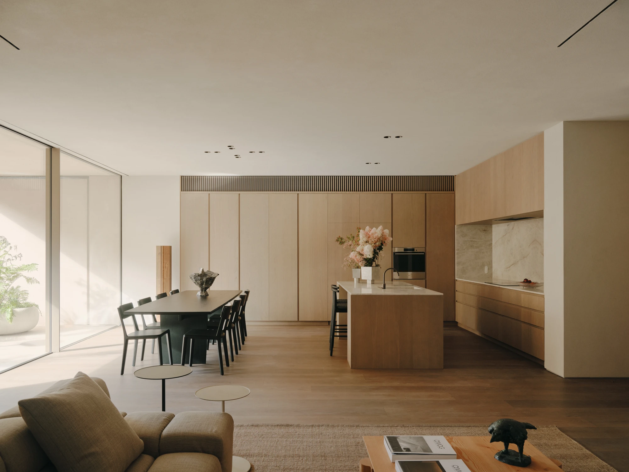 49 Walsh Street by MORQ, photographed by Tom Ross — A kitchen island in pale timber stands between the dining table and a stone splashback, with the living room and glazed wall to the left.