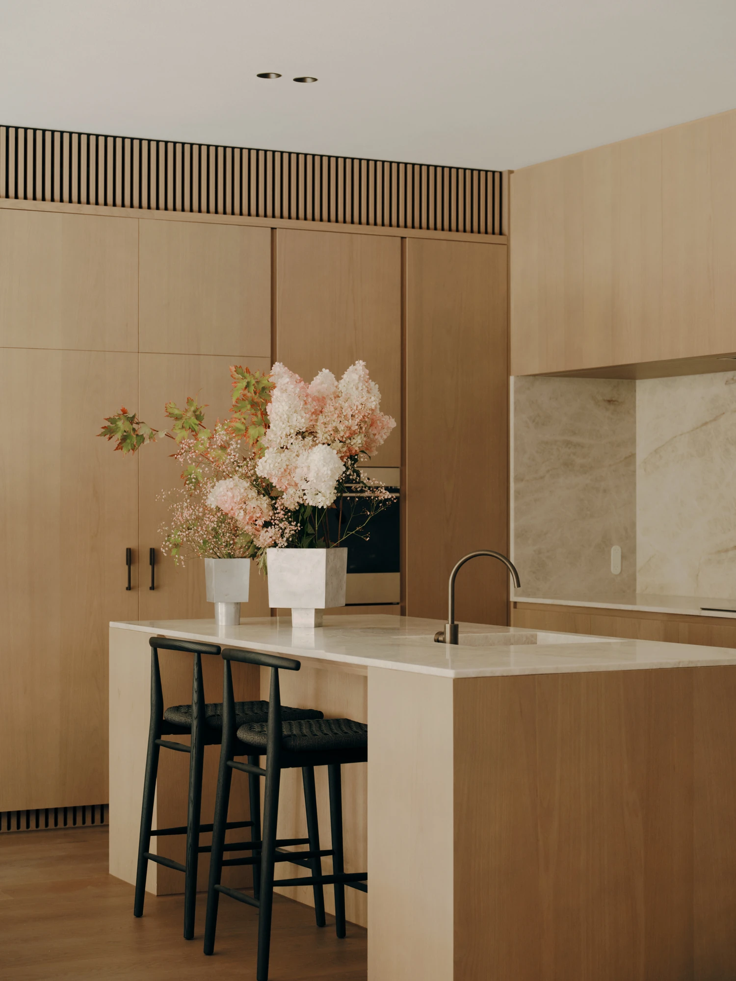 49 Walsh Street by MORQ, photographed by Tom Ross — The kitchen island in close view: pale timber carcass, stone benchtop, black tapware and two black woven bar stools, with hydrangeas in stone vessels and a slatted timber band above the cabinetry.