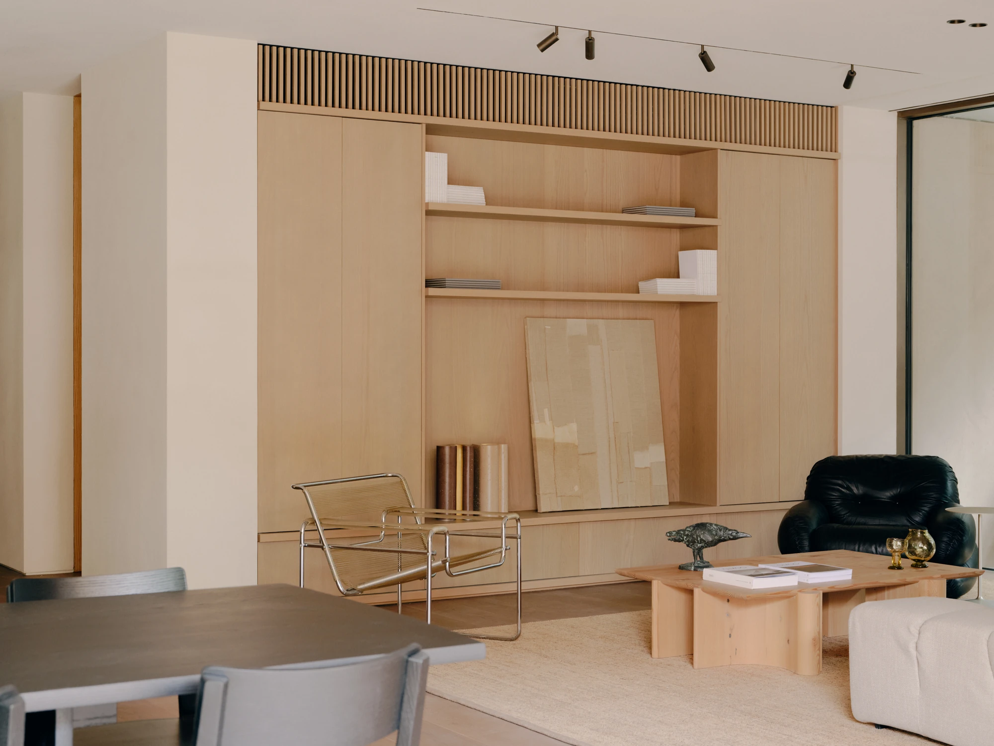 49 Walsh Street by MORQ, photographed by Tom Ross — A living room joinery wall in pale timber, with open shelves, a pale collaged panel leaning at floor level and bronze cylinders alongside.