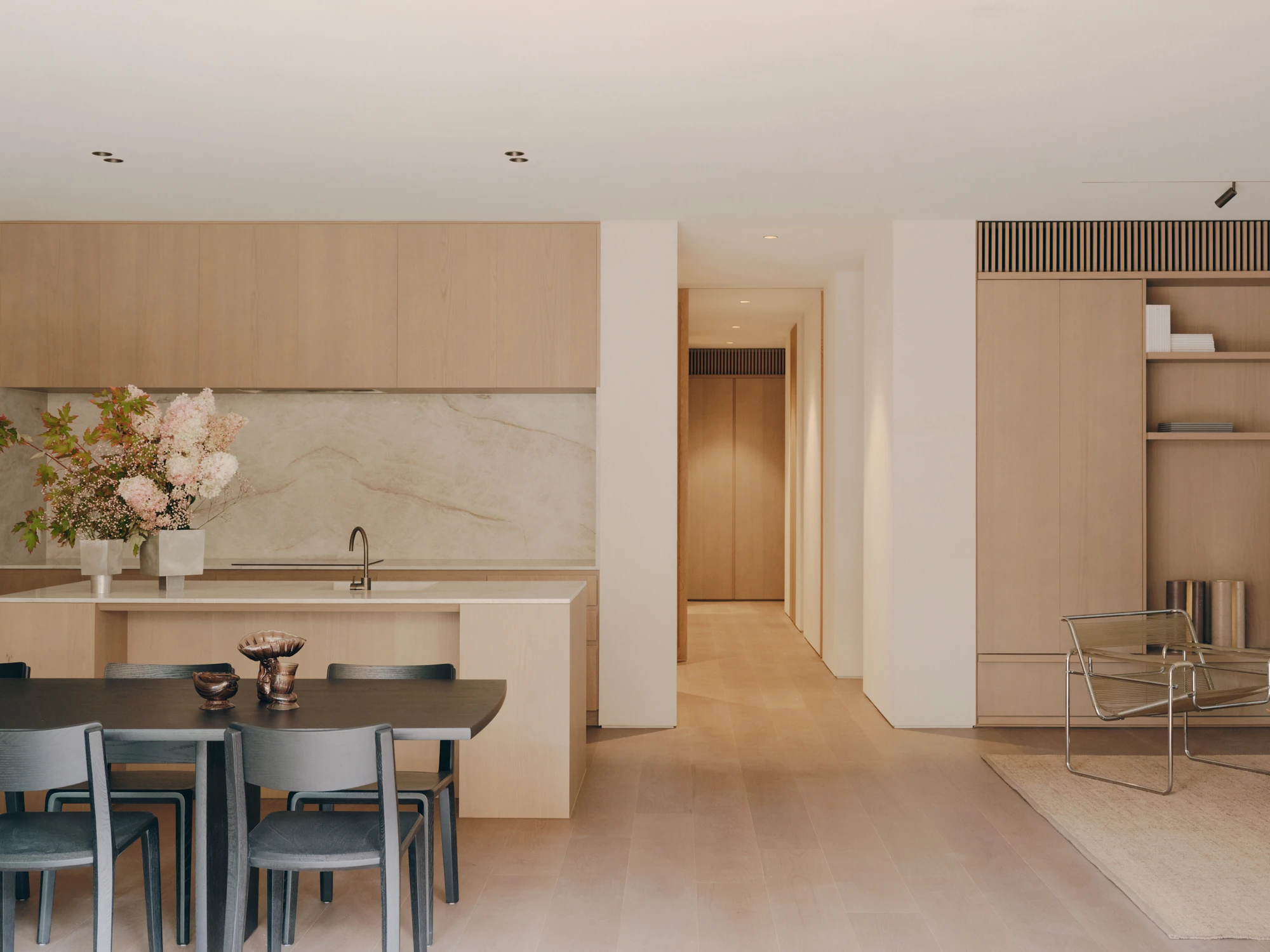 49 Walsh Street by MORQ, photographed by Tom Ross — The kitchen bench and stone splashback seen from the dining table, with a hallway of full-height doors running away to the right and a tubular steel chair on a pale rug.