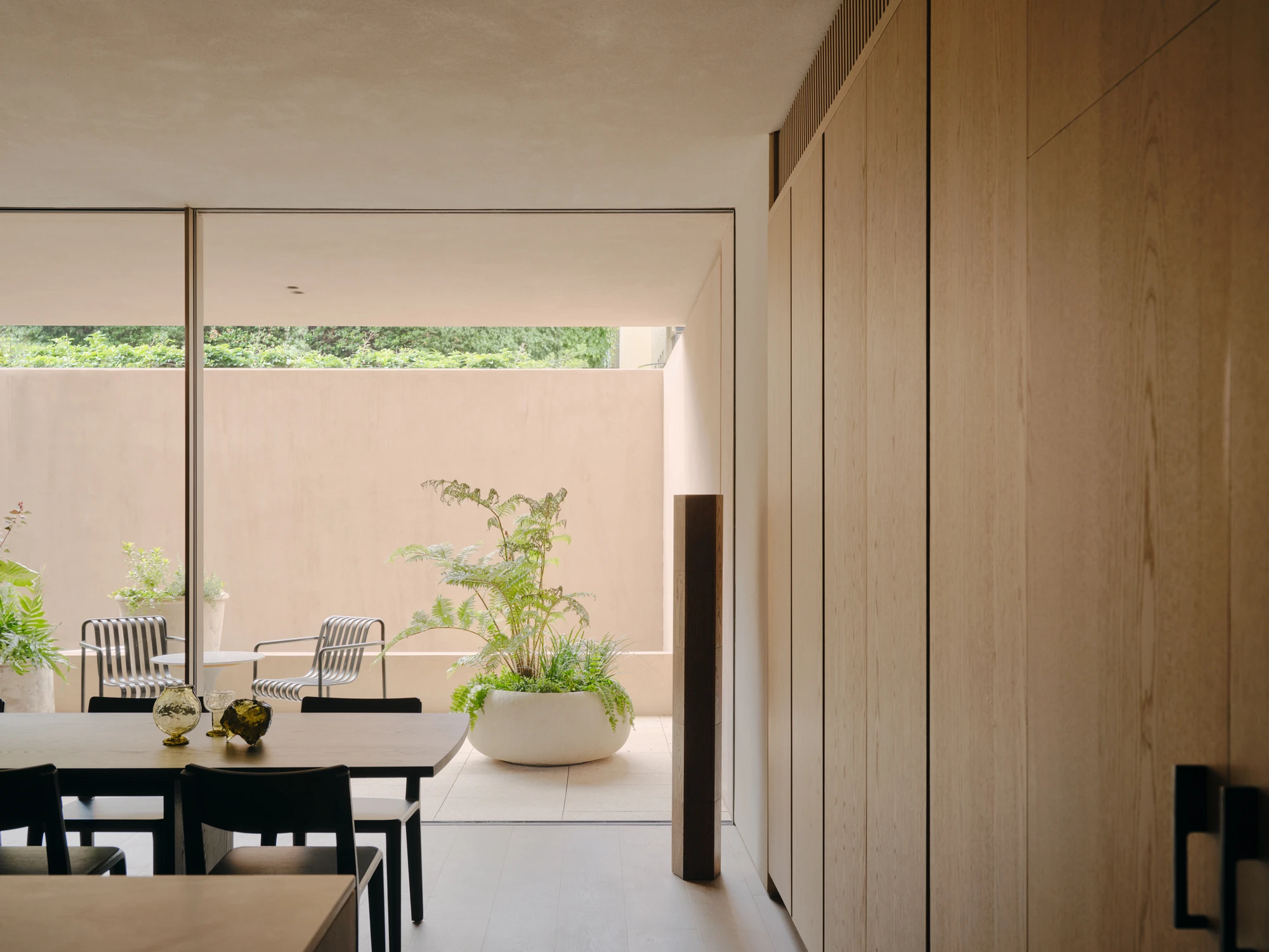 49 Walsh Street by MORQ, photographed by Tom Ross — Looking out through full-height glazing to a terrace where a fern sits in a round planter and striped chairs face the render wall, with tree canopy visible above it.