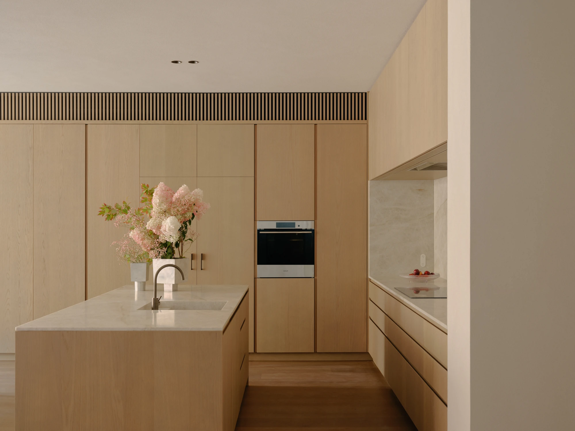 49 Walsh Street by MORQ, photographed by Tom Ross — A kitchen island in pale timber with a stone benchtop and integrated sink, backed by cabinetry with an integrated oven and a slatted band running above.