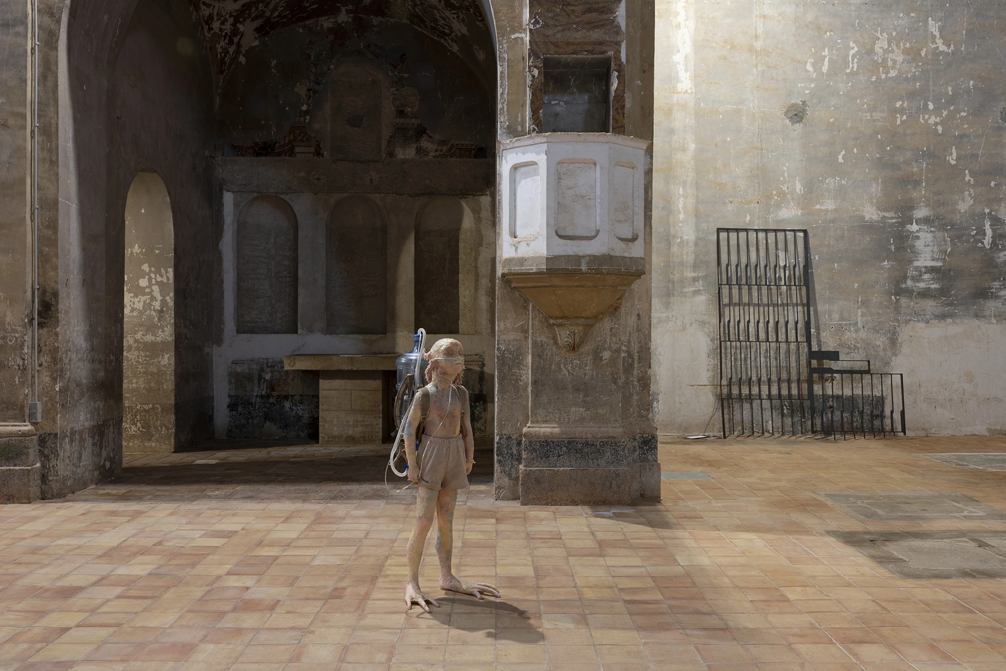 KIN TOO by Spiritvessel, photographed by Roberto Ruiz — Michele Gabriele's July 2nd, a life-size silicone figure carrying a water bottle on its back, standing beneath the church's white pulpit near Jordi Mitjà's leaning steel grille.