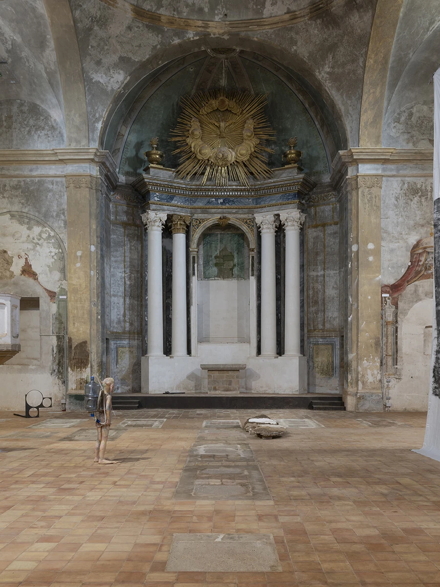 KIN TOO by Spiritvessel, photographed by Roberto Ruiz — A wide view down the nave of the disused Church of Sant Feliu in Lladó, its altar crowned by a gilt sunburst, with Michele Gabriele's silicone figure on the terracotta floor and Andrew Birk's…