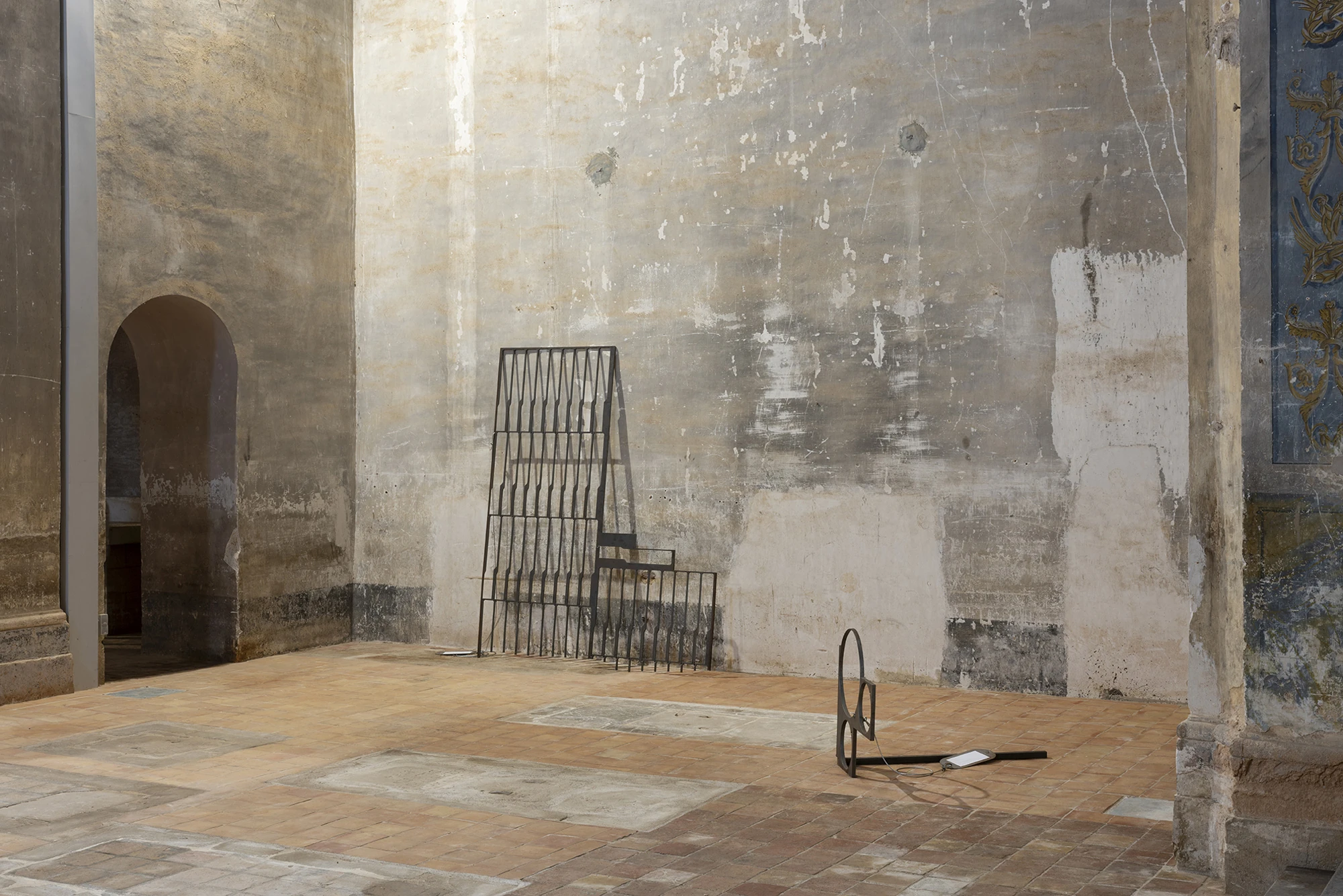 KIN TOO by Spiritvessel, photographed by Roberto Ruiz — Two works by Jordi Mitjà against a bare plaster wall: a tall steel grille and a low form of interlocking cut circles trailing a steel cable and a pale metal tag.