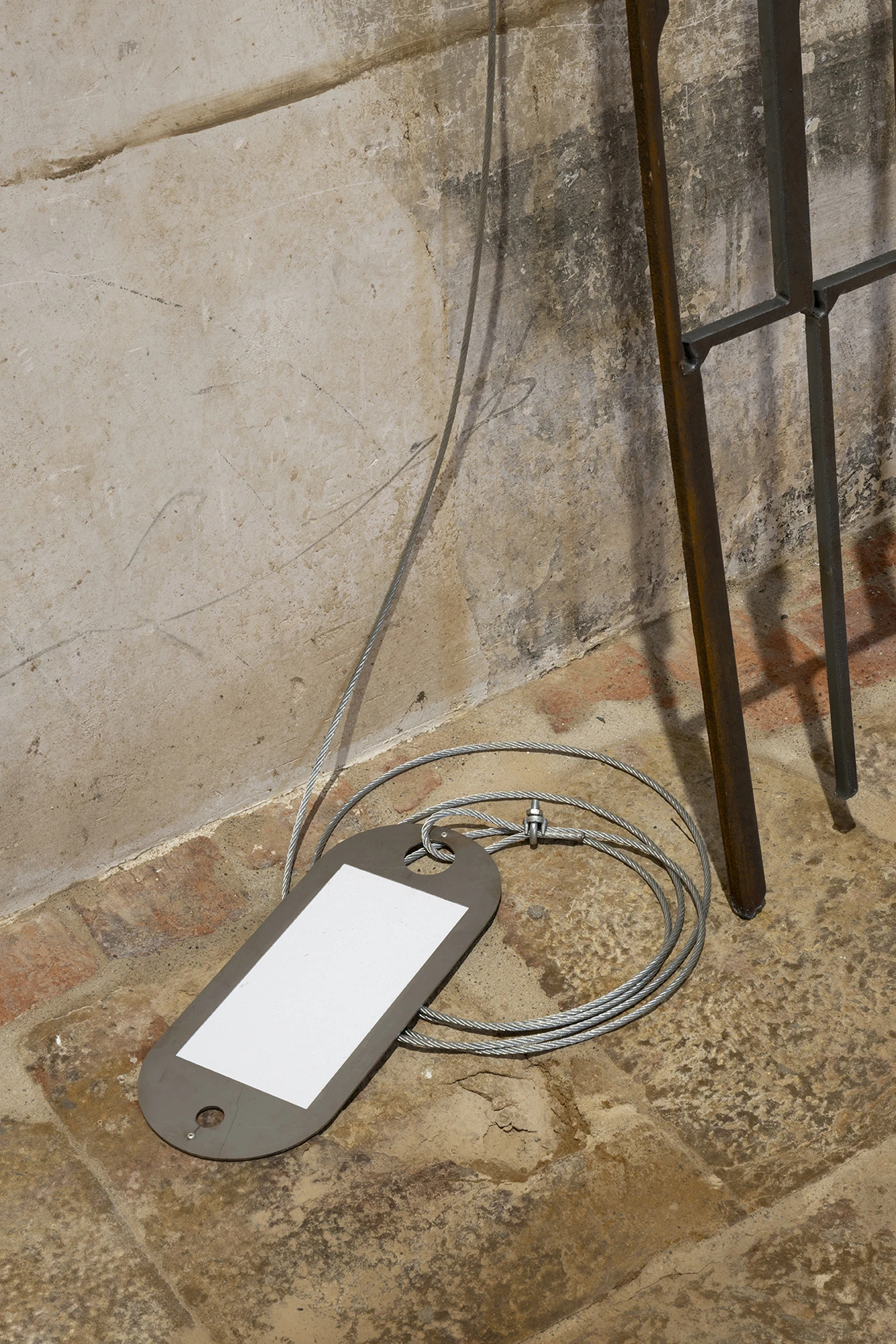KIN TOO by Spiritvessel, photographed by Roberto Ruiz — Detail of Jordi Mitjà's work, a steel dog-tag plate with a blank white field and a coiled steel cable resting on the stone floor beside an iron leg.
