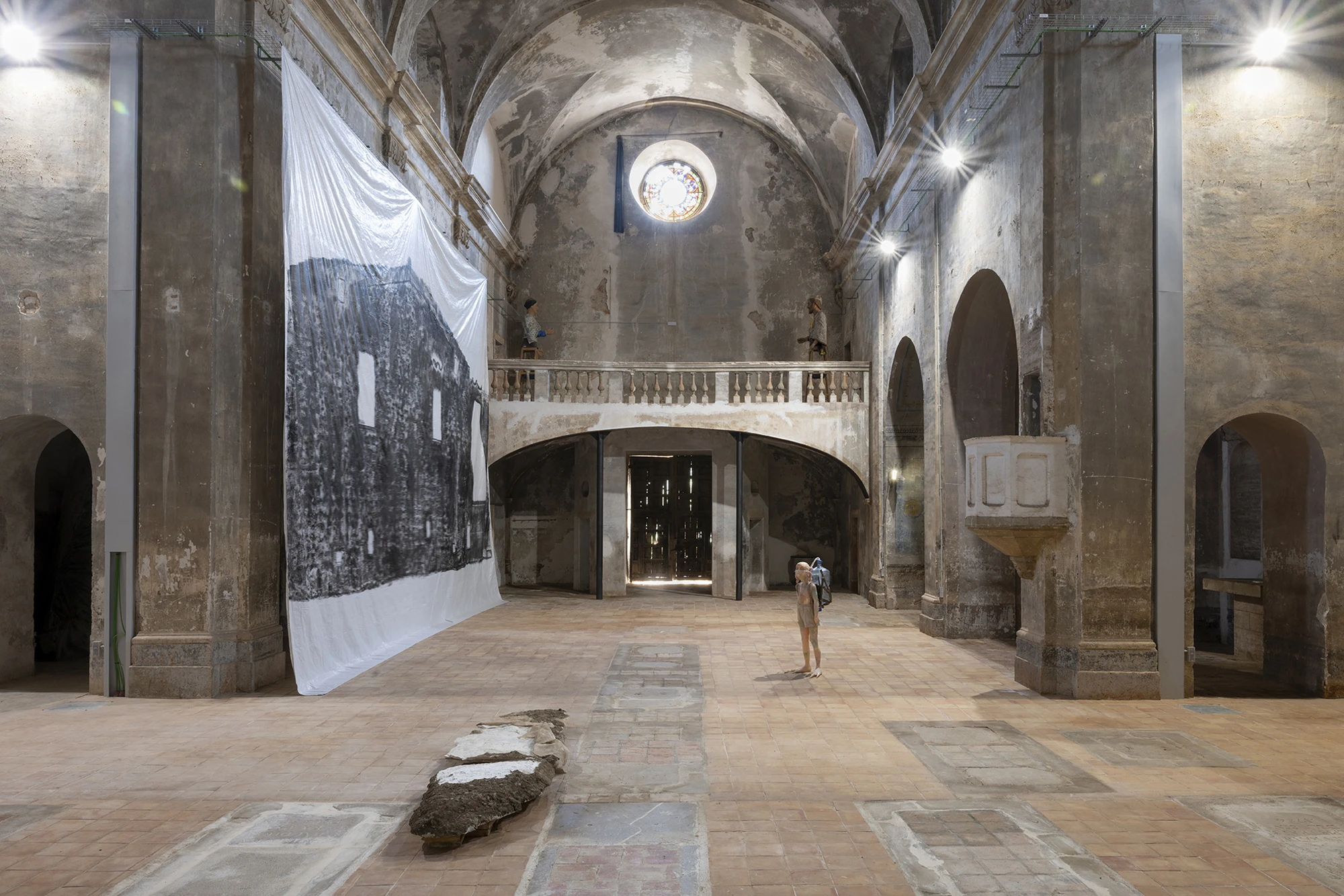 KIN TOO by Spiritvessel, photographed by Roberto Ruiz — The nave of Sant Feliu with Andrew Birk's Floating House hung between piers, Michele Gabriele's figure on the floor, and a plaster Ghost slab in the foreground under the rose window.